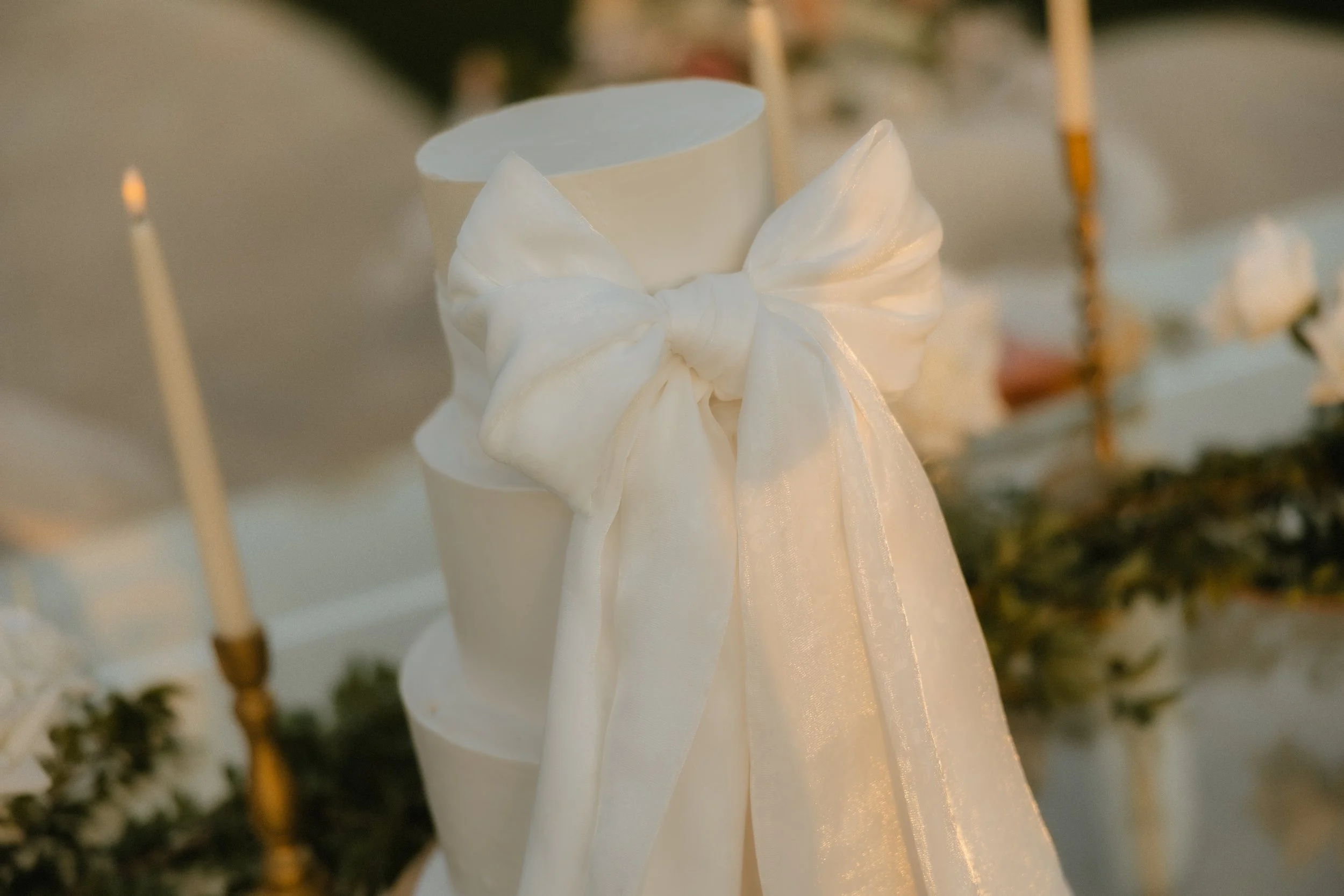 Elegant white cake with large bow decoration, surrounded by candles and greenery.