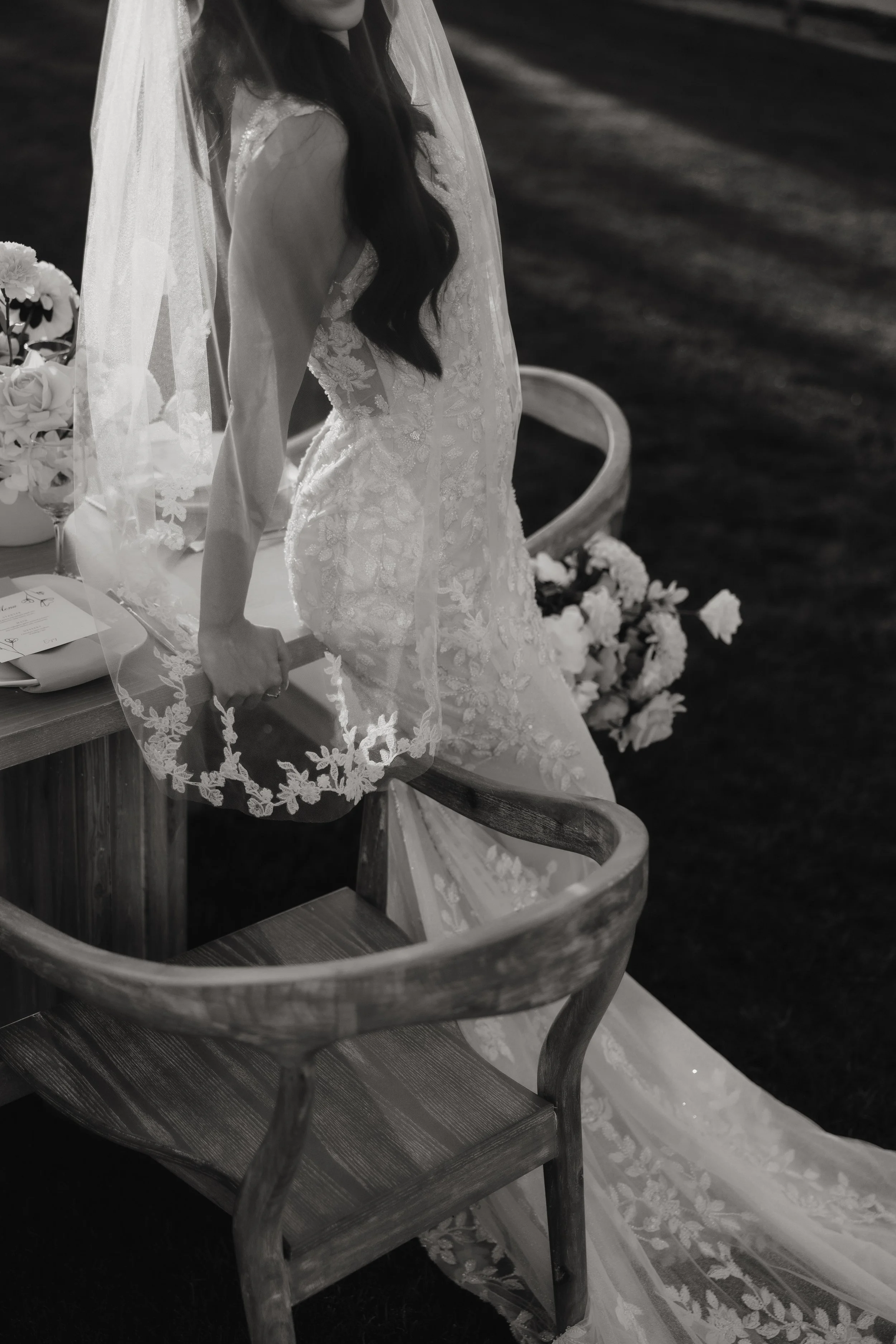 Bride in lace wedding dress and veil sitting on wooden chair outdoors