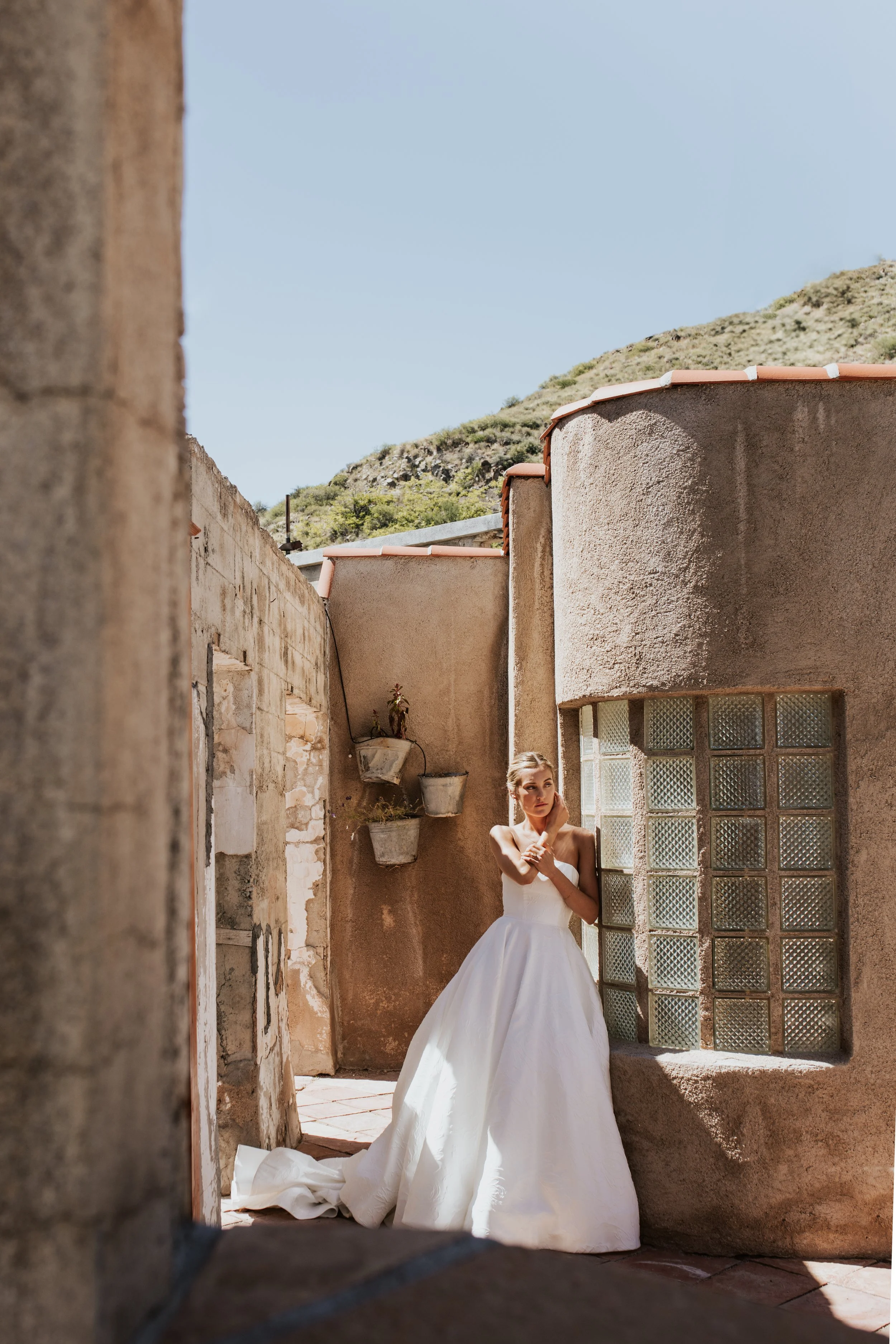 Bride in white gown standing against rustic wall with glass blocks and potted plants, outdoor setting, sunny day.