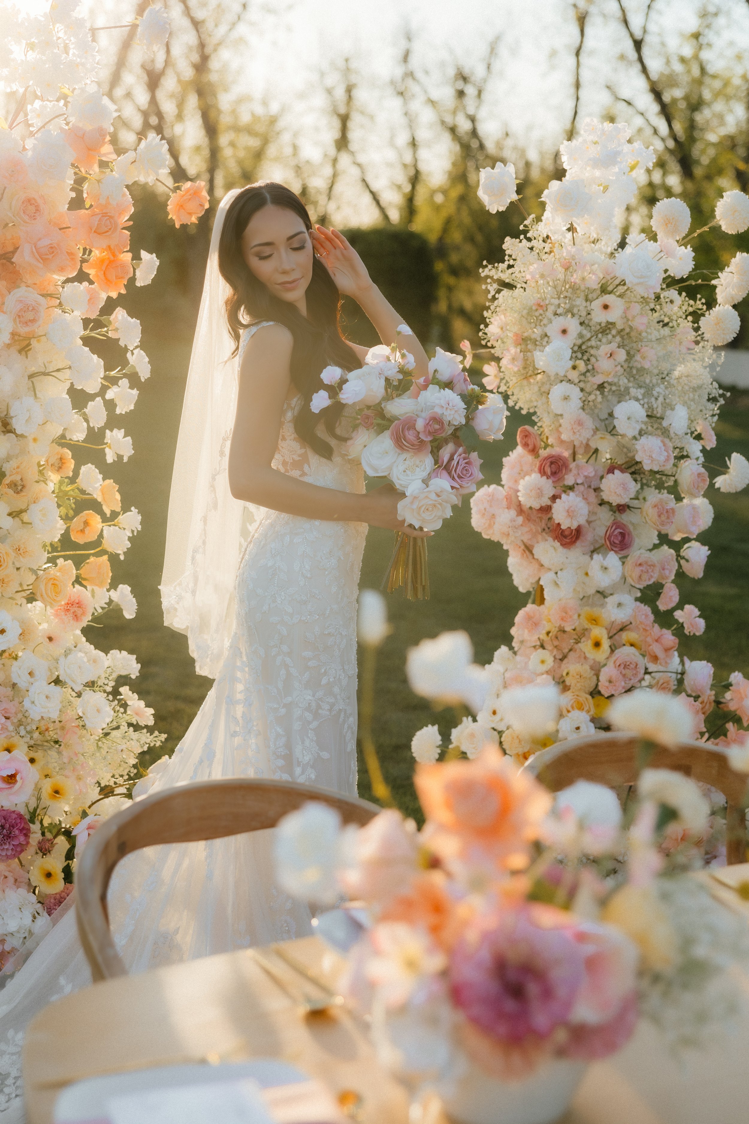 Bride in white lace dress holding bouquet surrounded by colorful floral arrangements outdoors