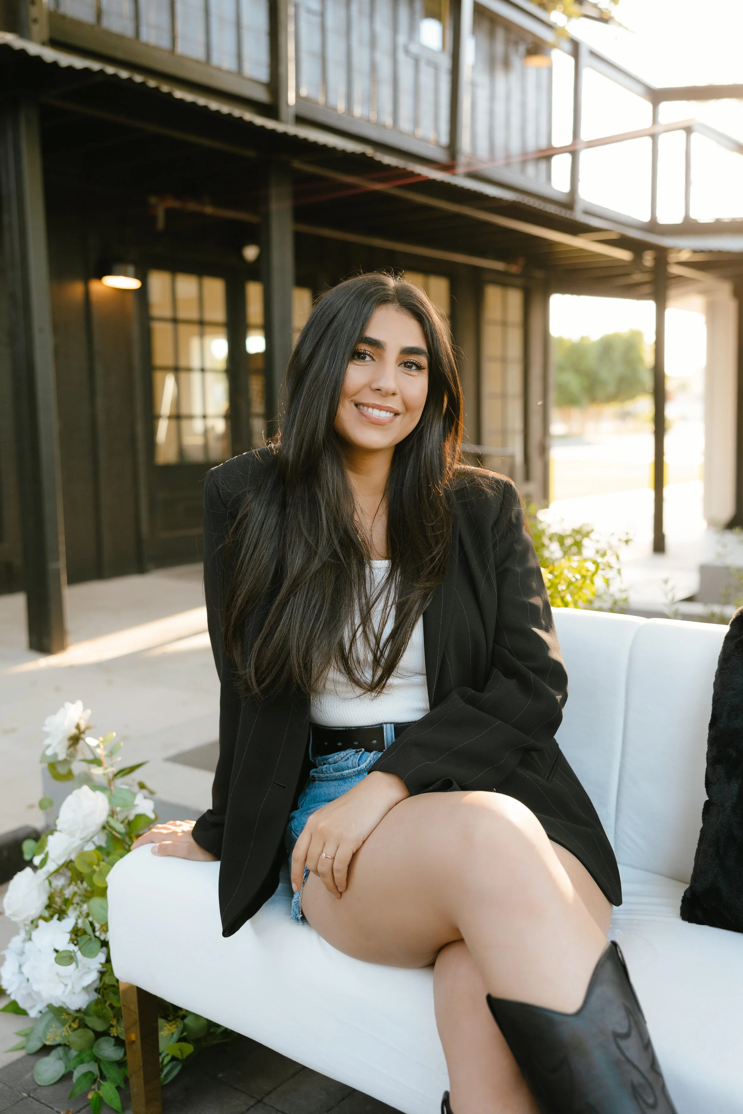 Woman sitting on white couch outdoors, wearing black blazer, white top, denim shorts, black boots, with long dark hair, smiling, in front of building.