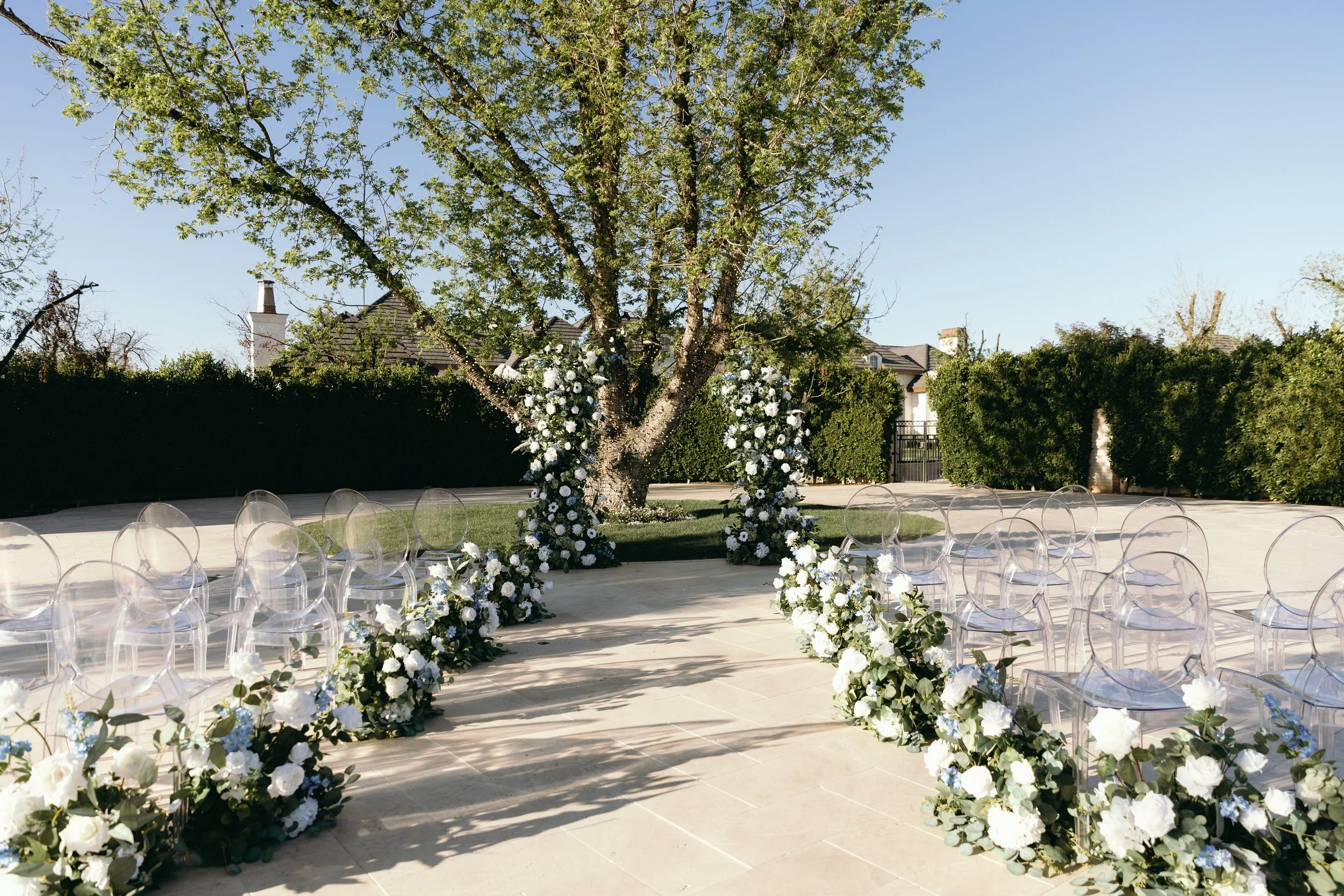 Outdoor wedding ceremony setup with clear chairs and floral arrangements, featuring white and blue flowers under a large tree.