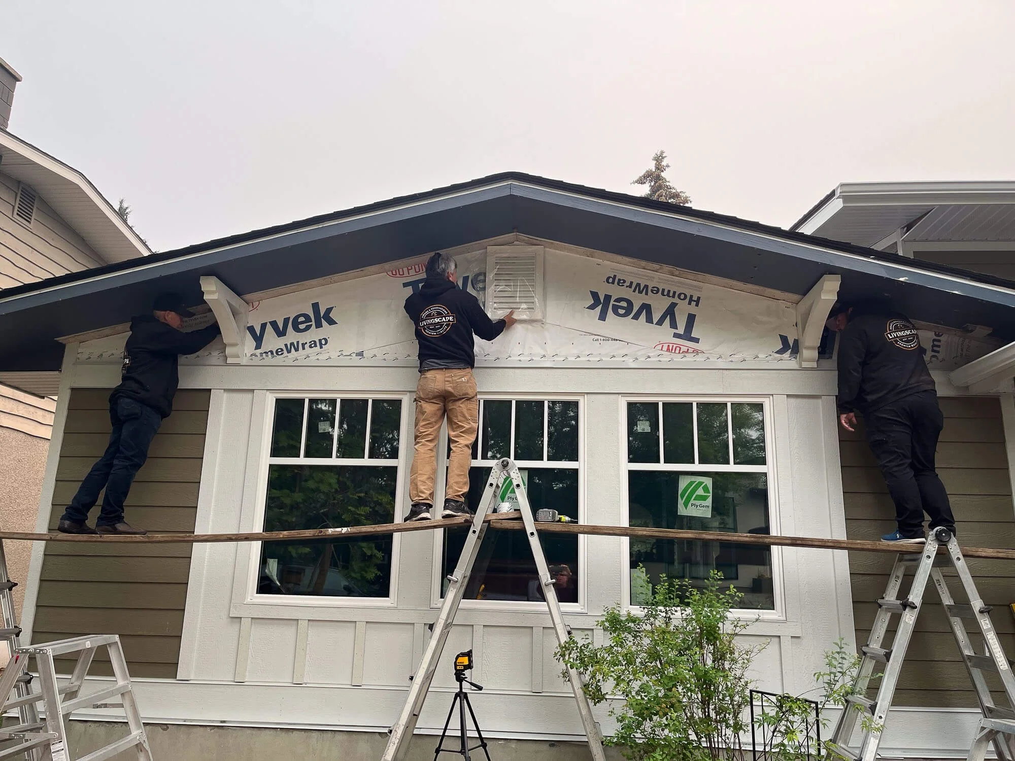Three siding contractors calgary installing or repairing siding and trim on the exterior of a house under a cloudy sky, using ladders and a wooden plank for support.