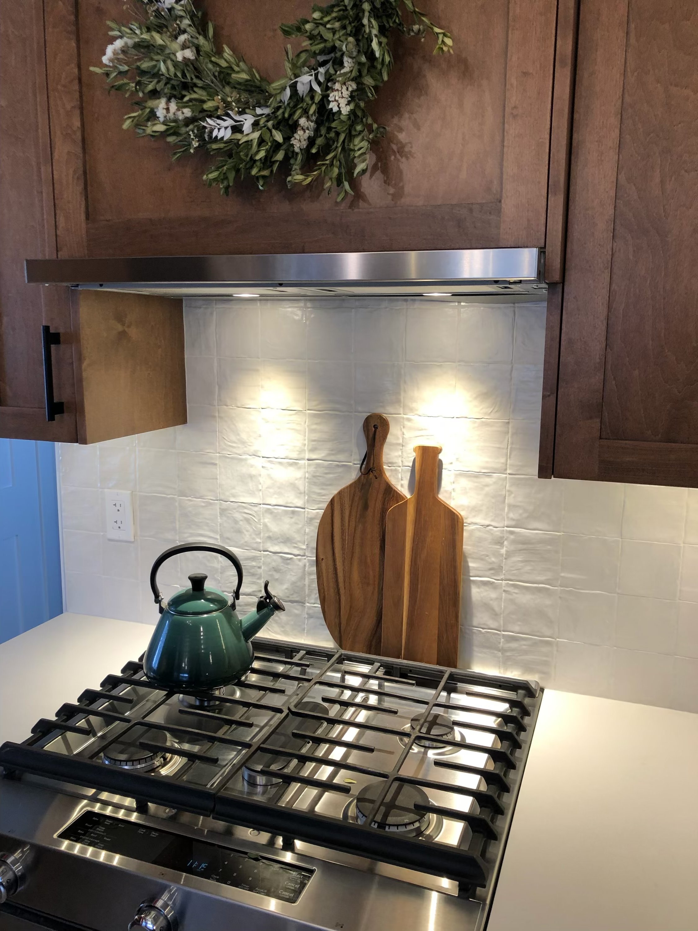 Green kettle on a stainless steel gas stove, with two wooden cutting boards behind it, mounted on a white tiled backsplash in a kitchen.