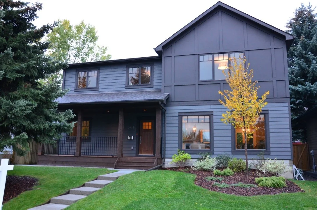 A two-story blue house with a front porch, a small yard with green grass, a young tree with yellow leaves, and some shrubs, with a wooden fence on the right side.