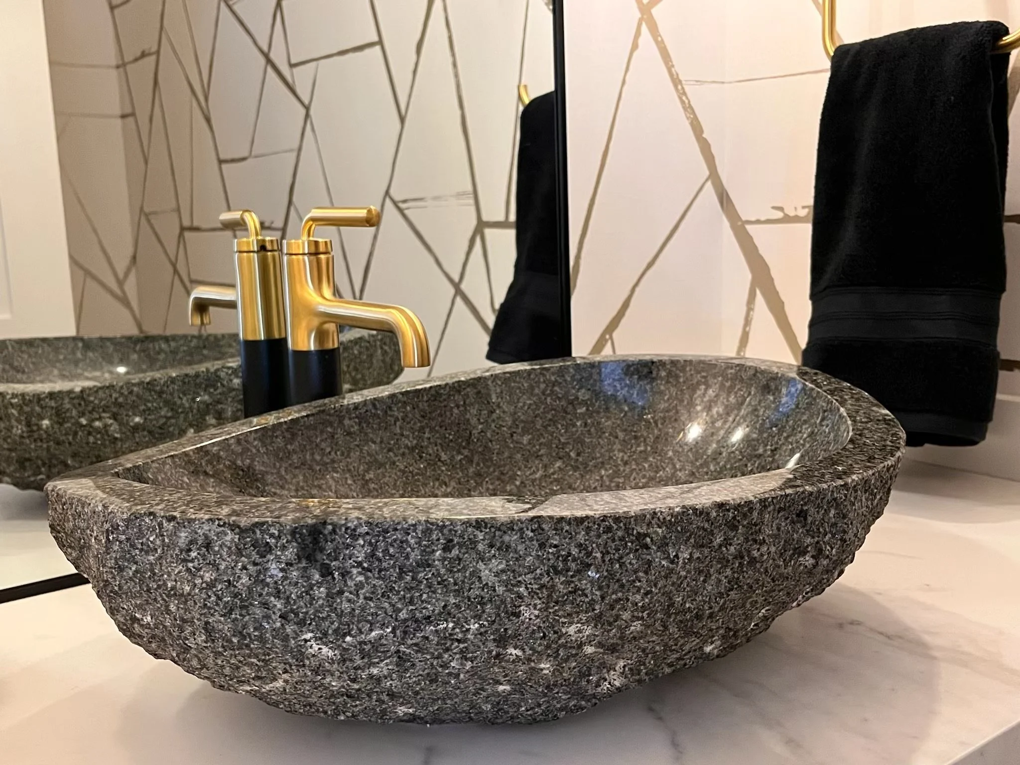 A granite bathroom sink with a gold faucet on a marble countertop, with a geometric pattern wall and a black towel hanging on a gold towel rack in the background.