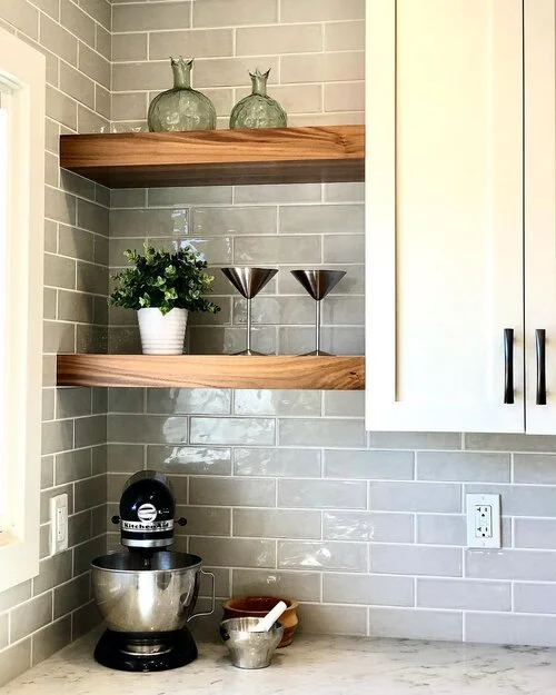 Kitchen corner with two wooden floating shelves holding decorative glass vases, a potted plant, and two metallic glasses, alongside a coffee machine and a bowl on a marble countertop.
