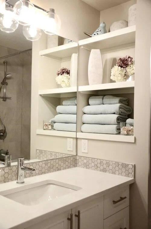 Bathroom vanity with stacked towels, decorative vases, and shelves with flowers and decor, reflected in a mirror.