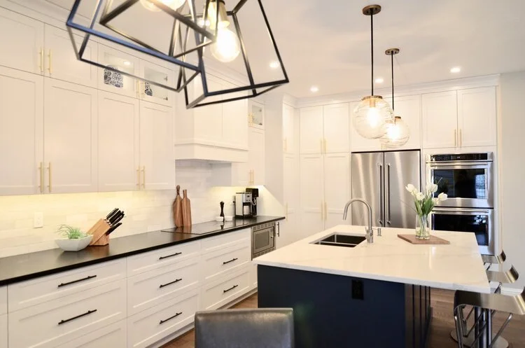 Modern white kitchen with black countertop, large island with sink and flowers, stainless steel appliances, and stylish black light fixtures.