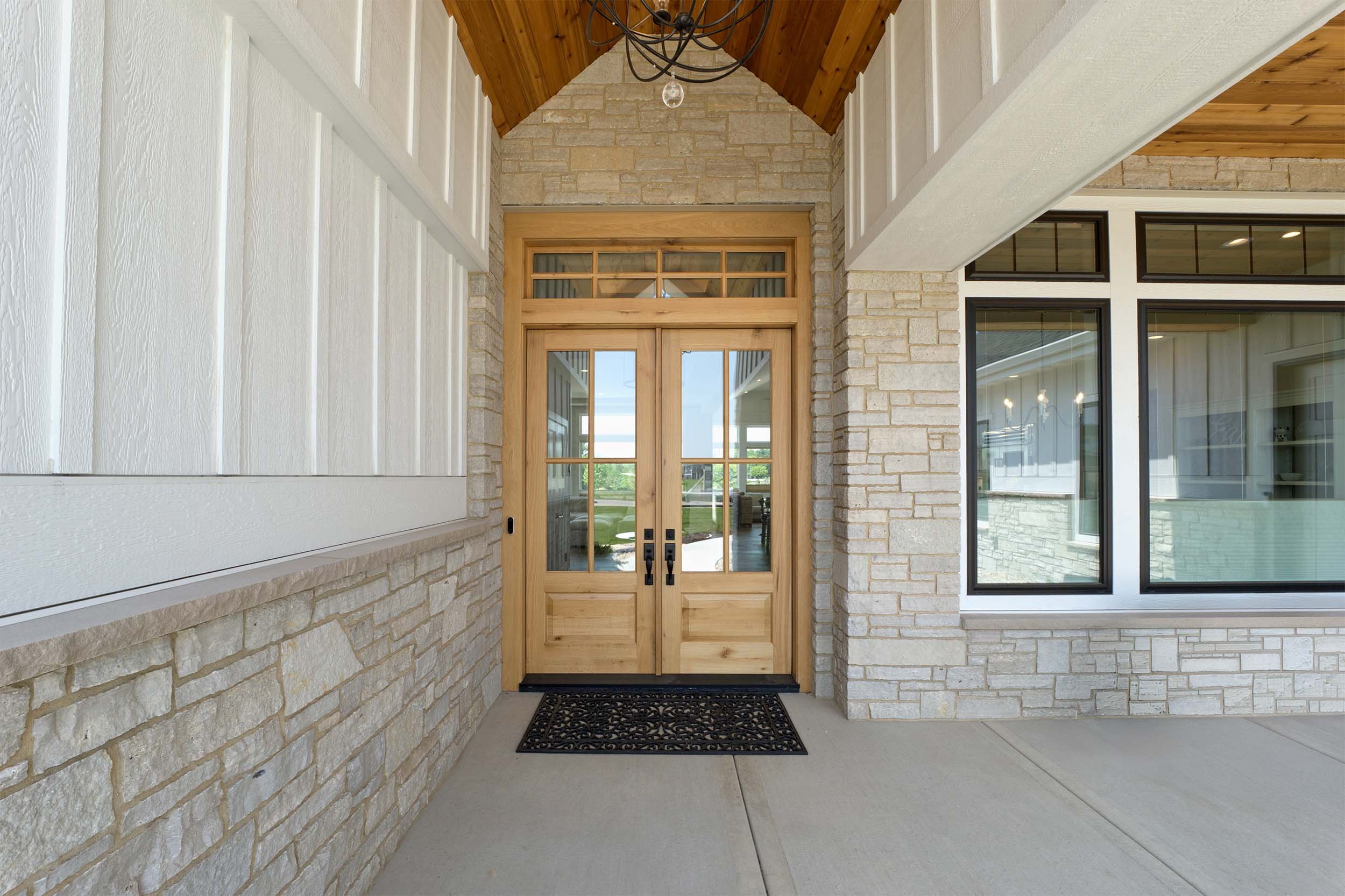 Front porch with wooden door, stone and white siding walls, large window, and a decorative chandelier overhead.