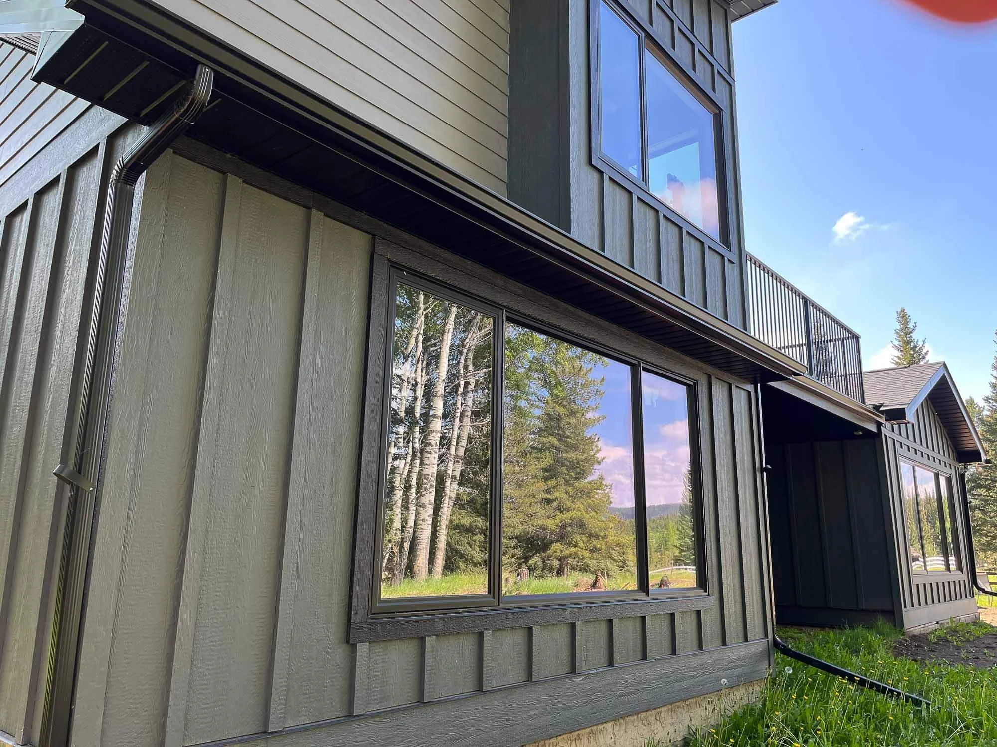 Close-up of a modern house exterior with gray wood siding, large windows reflecting trees and sky, and a metal gutter system. The house has a two-story structure with a balcony on the upper level and surrounded by greenery.