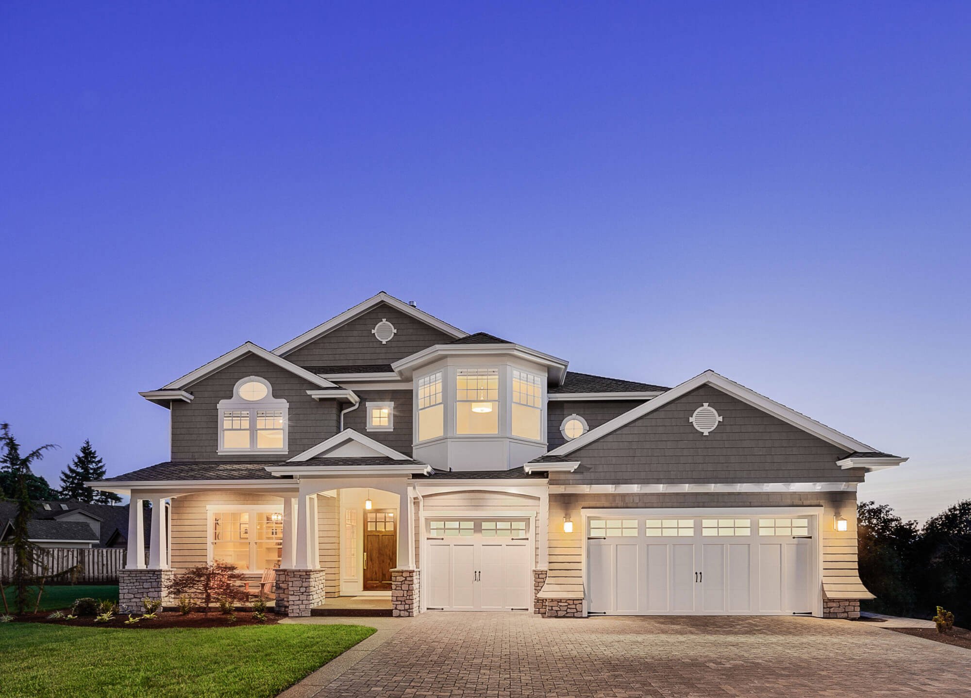 A two-story house with a gray and white exterior, front porch, large windows, and a three-car garage, illuminated by exterior lights during dusk.