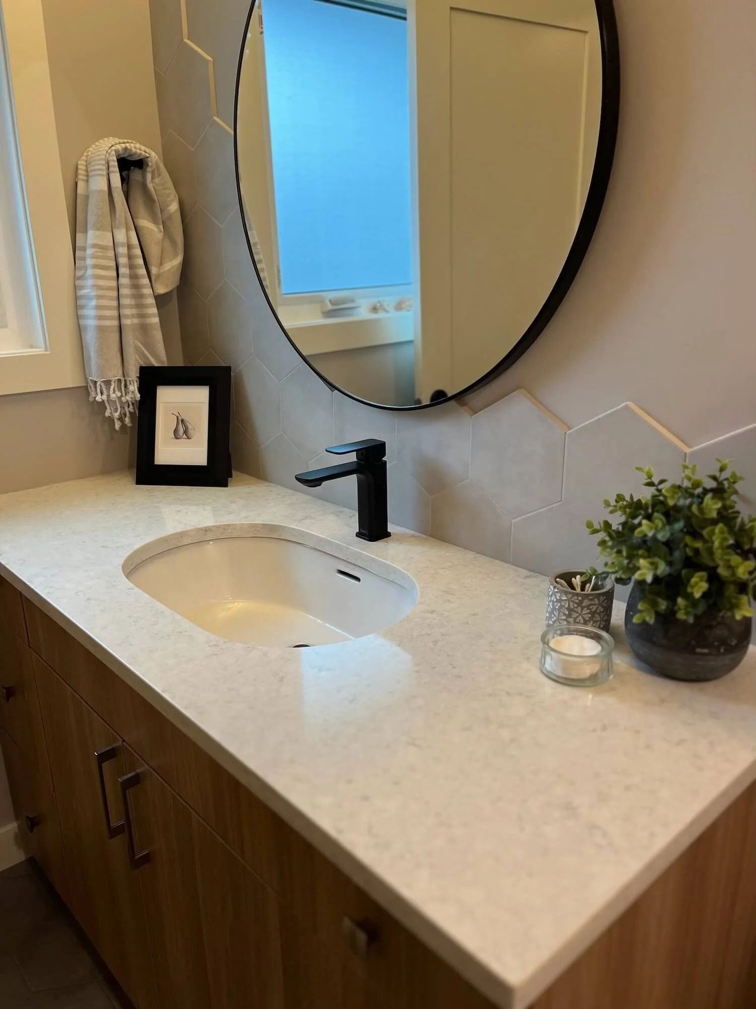 Bathroom vanity with a white countertop, black faucet, oval mirror, and decorative items including a framed picture, a small potted plant, and candles.