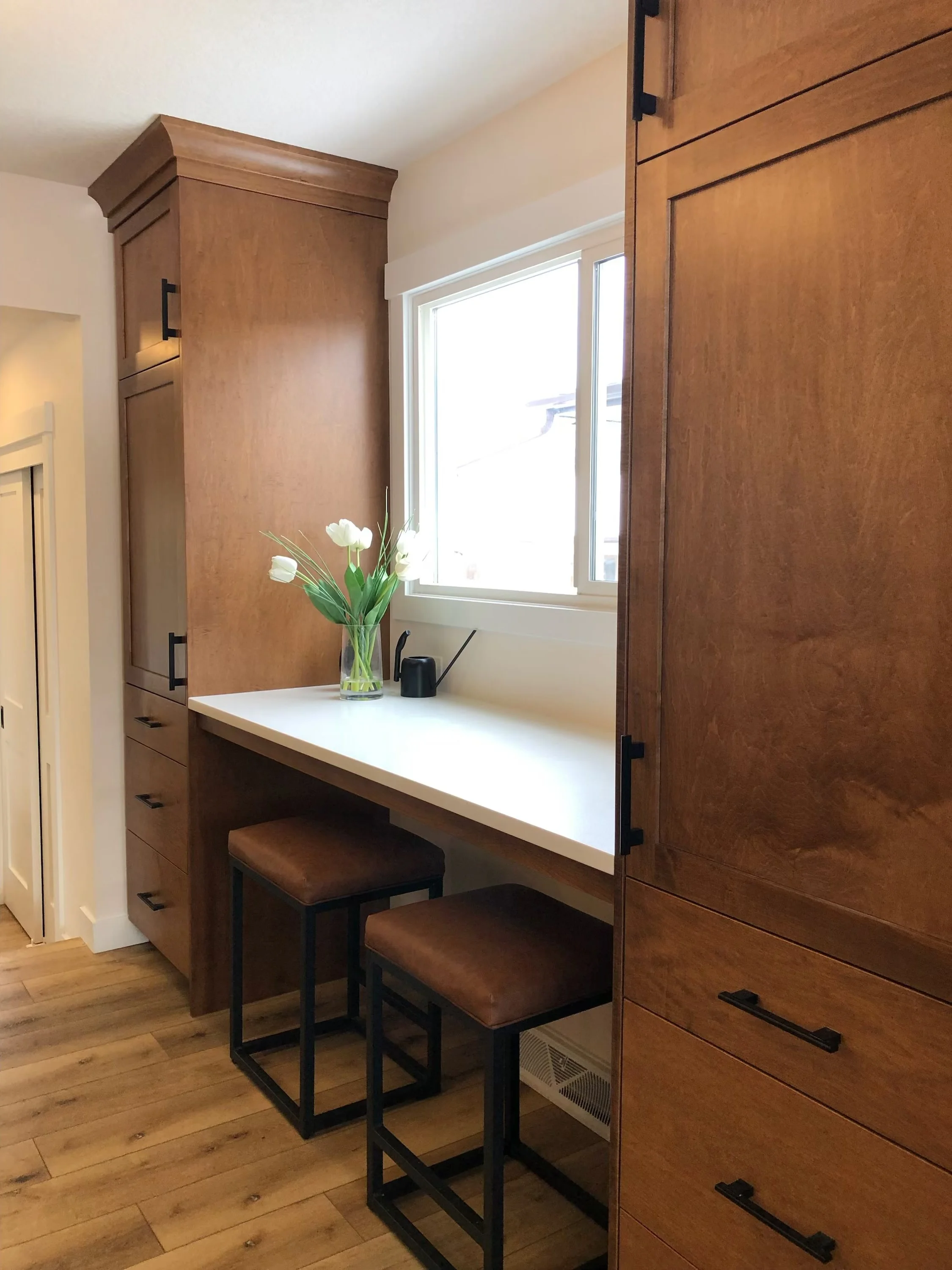A kitchen corner with a white countertop under a window, featuring a vase with white tulips and an electric kettle. There are two brown cushioned stools tucked under the counter, surrounded by wooden cabinets and drawers.