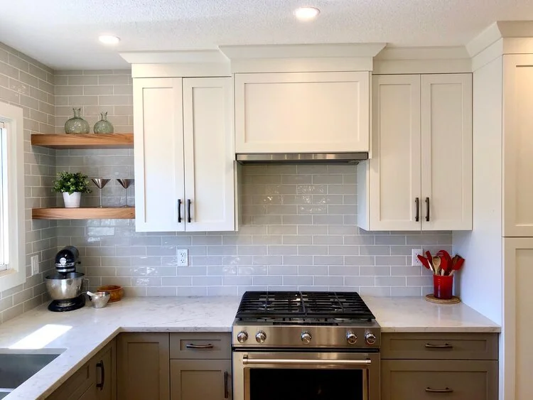 Modern kitchen with white cabinets, gray tile backsplash, and a gray lower cabinet. Items on the counter include a stand mixer and bowls on the left, and a utensil holder on the right. There are open wooden shelves with decorative vases and plant on 