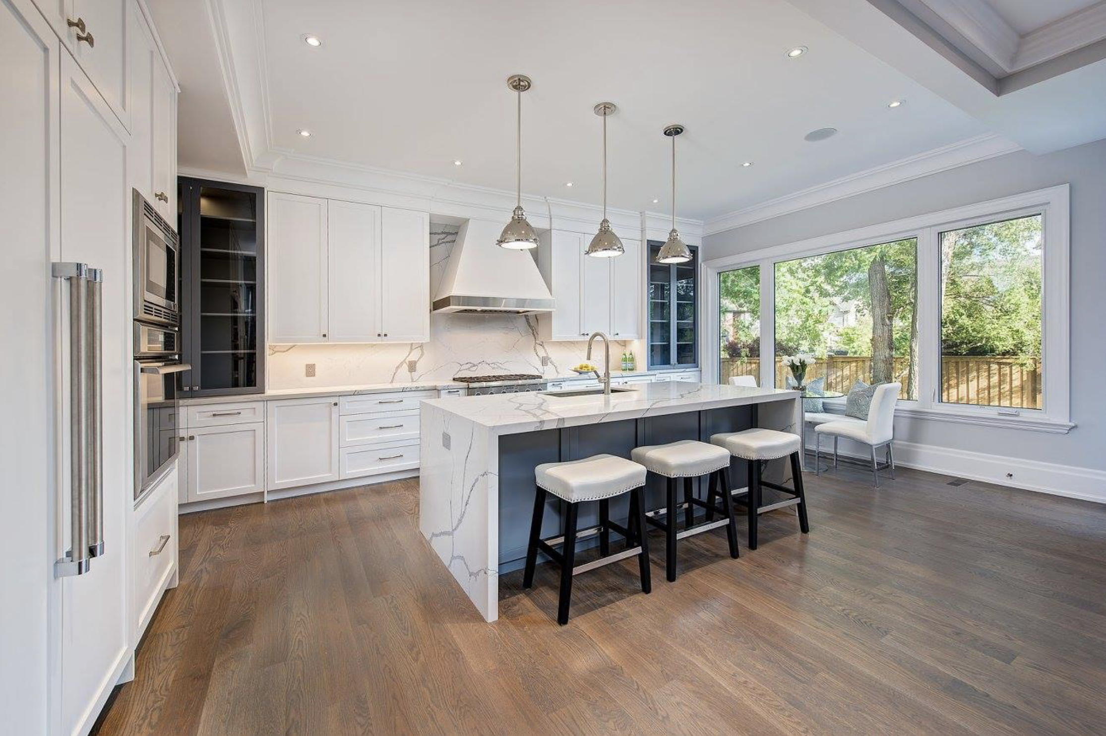 Modern kitchen with white cabinets, marble backsplash, central island with marble countertop, three pendant lights, large windows with views of green outdoor space, hardwood floor, seating area with white chairs.