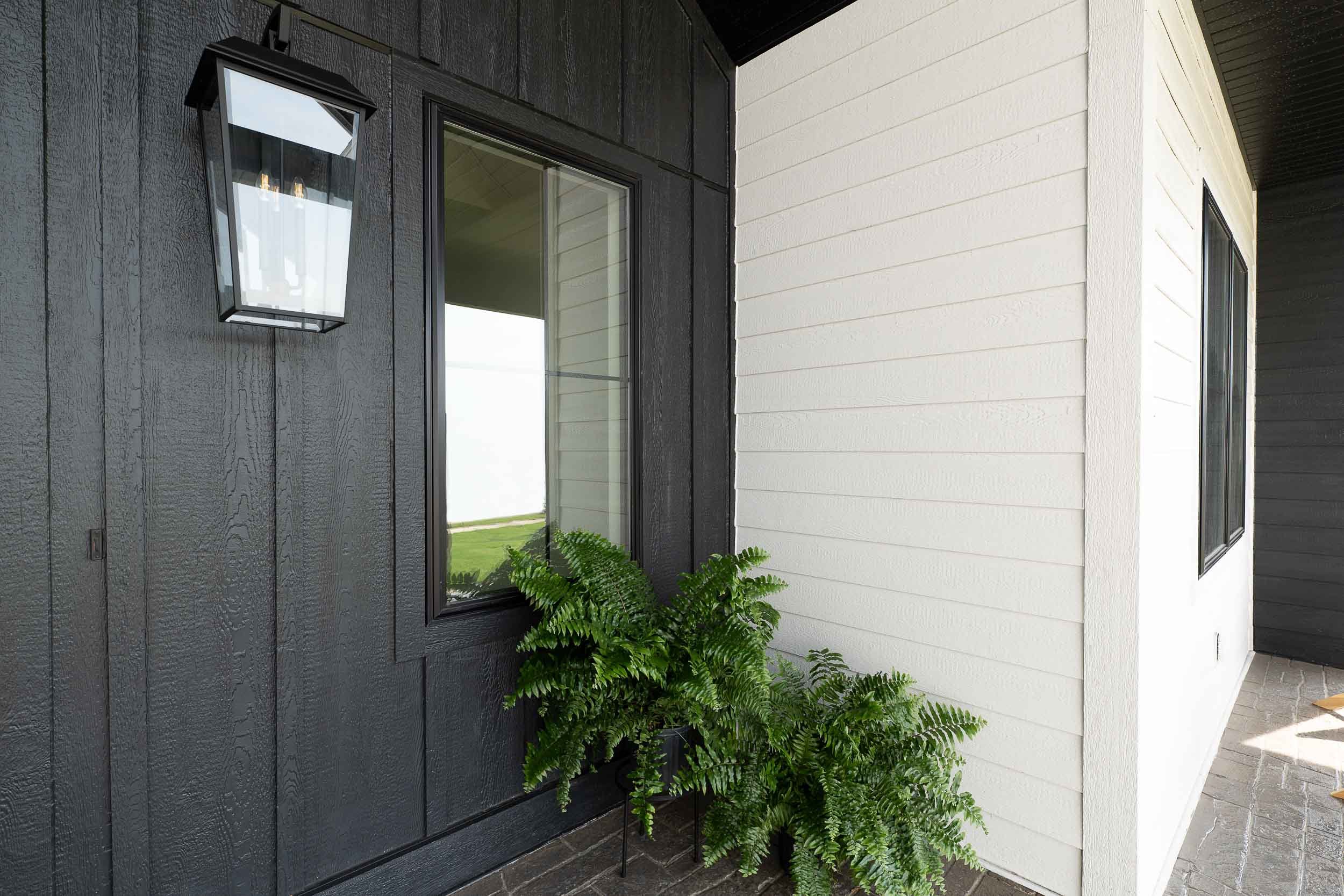 Exterior corner of modern house with black and white siding, black-framed window, black outdoor wall lantern, and green fern plant in a pot on the porch.