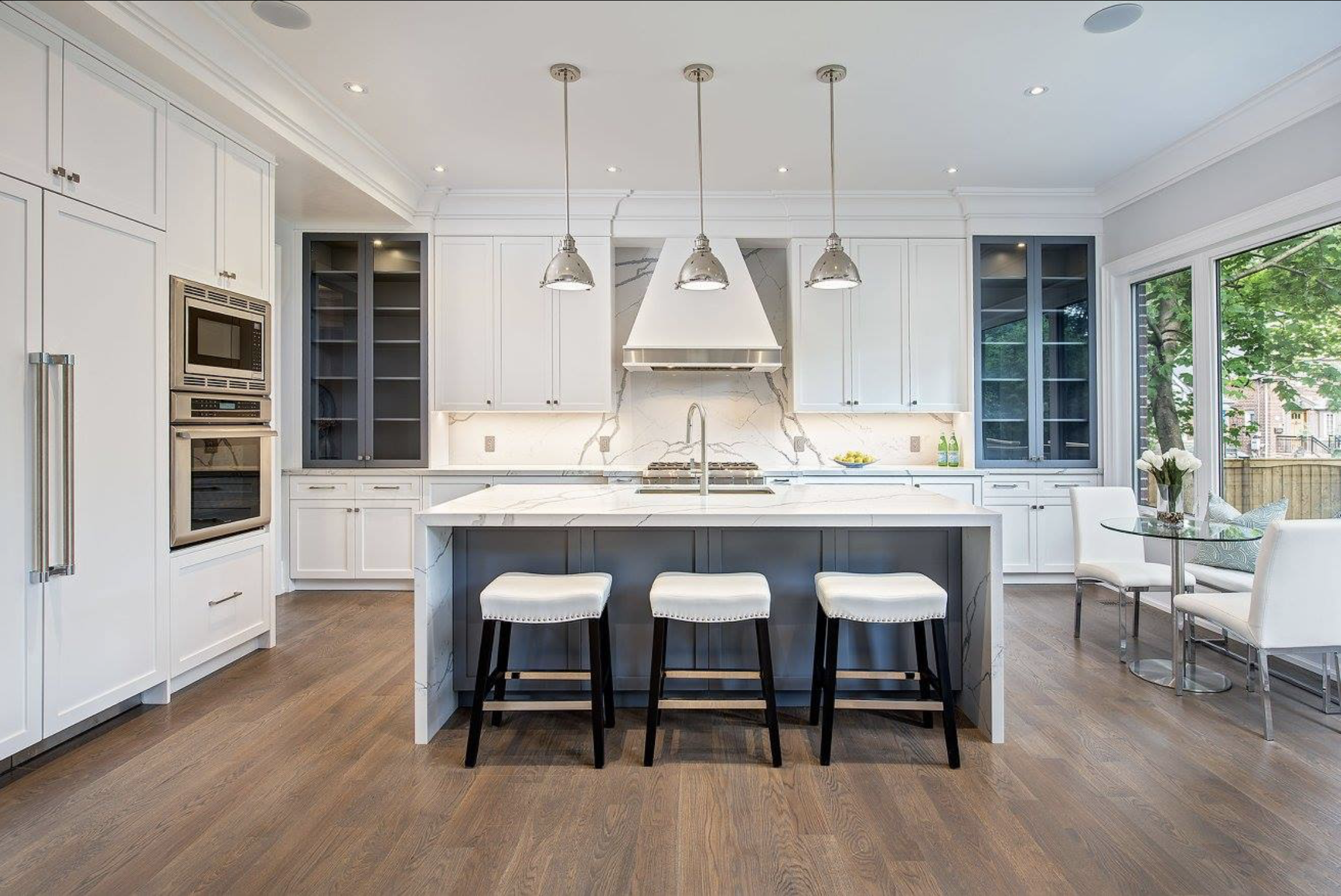Modern kitchen with white cabinets, marble island, three pendant lights, built-in appliances, and a dining area with white chairs near large windows showing greenery outside.