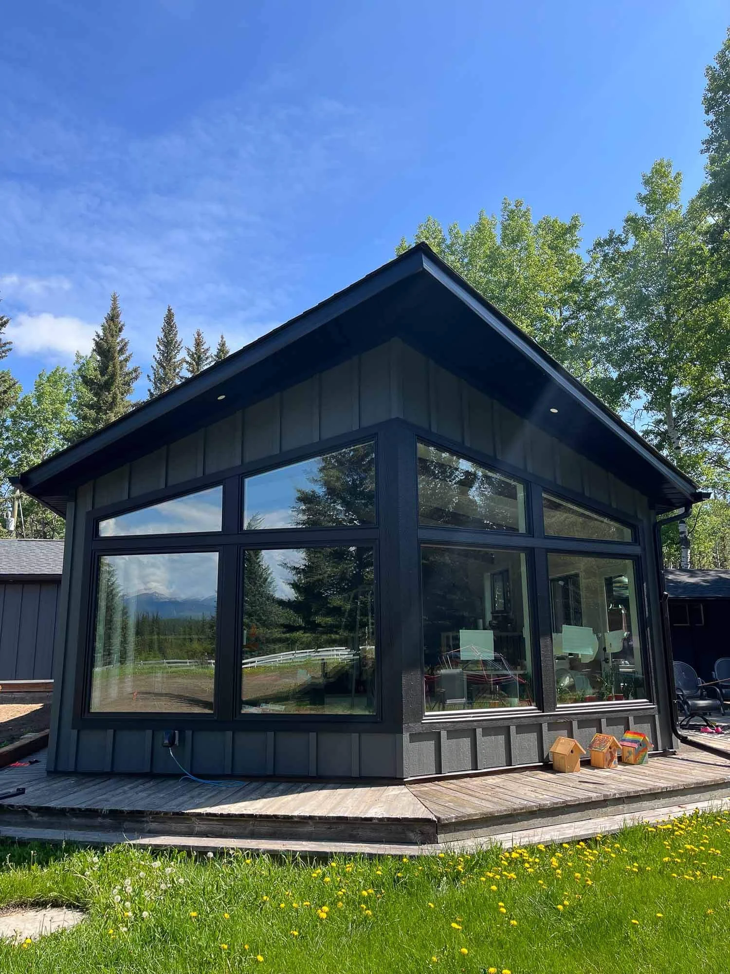 A modern house with large glass windows, black exterior trim, and a wooden deck surrounded by green grass and yellow flowers, set against a backdrop of trees and a blue sky.