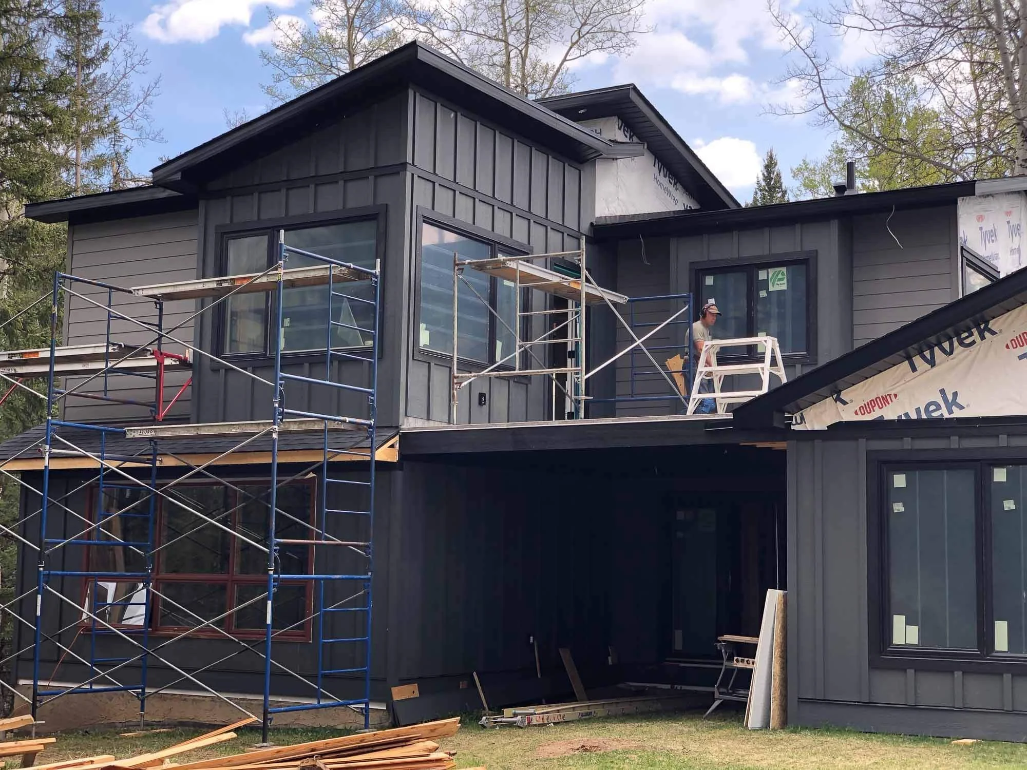 Construction workers installing black exterior siding on a modern two-story house with scaffolding set up on the front porch and upper level.