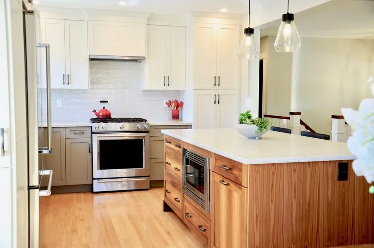 Modern kitchen with white cabinets, a beige kitchen island with a built-in microwave and drawers, a stainless steel stove with a red kettle on top, and pendant lights hanging above the island.