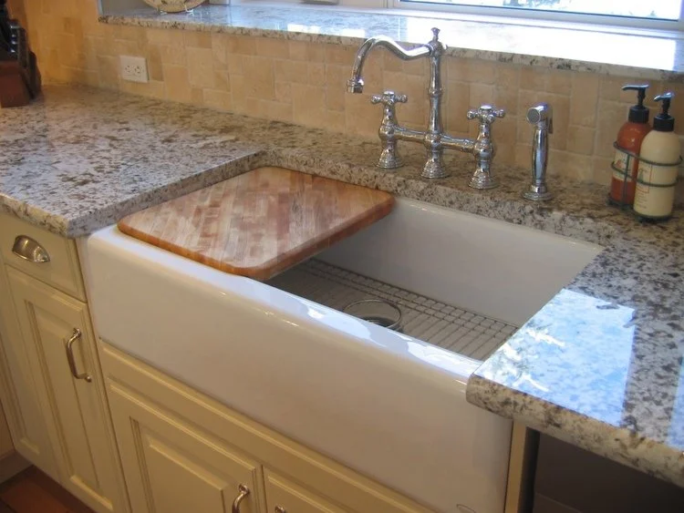Kitchen sink with a granite countertop, a wooden cutting board covering part of the sink, a vintage-style silver faucet, and soap dispensers. There is a window above the sink and cabinetry below.