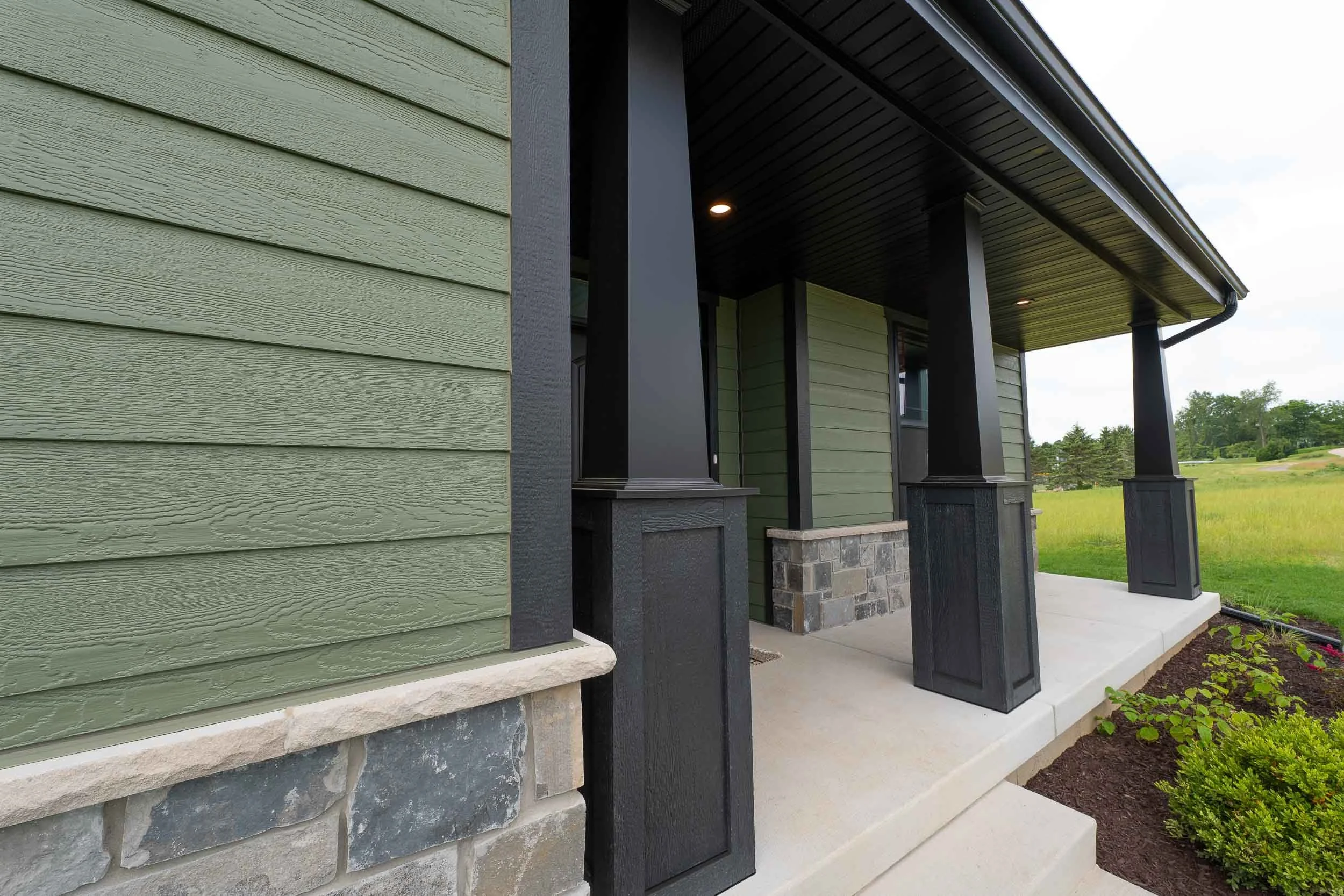Front porch of a house with green and black exterior siding, stone accents, a concrete porch, and garden plants.
