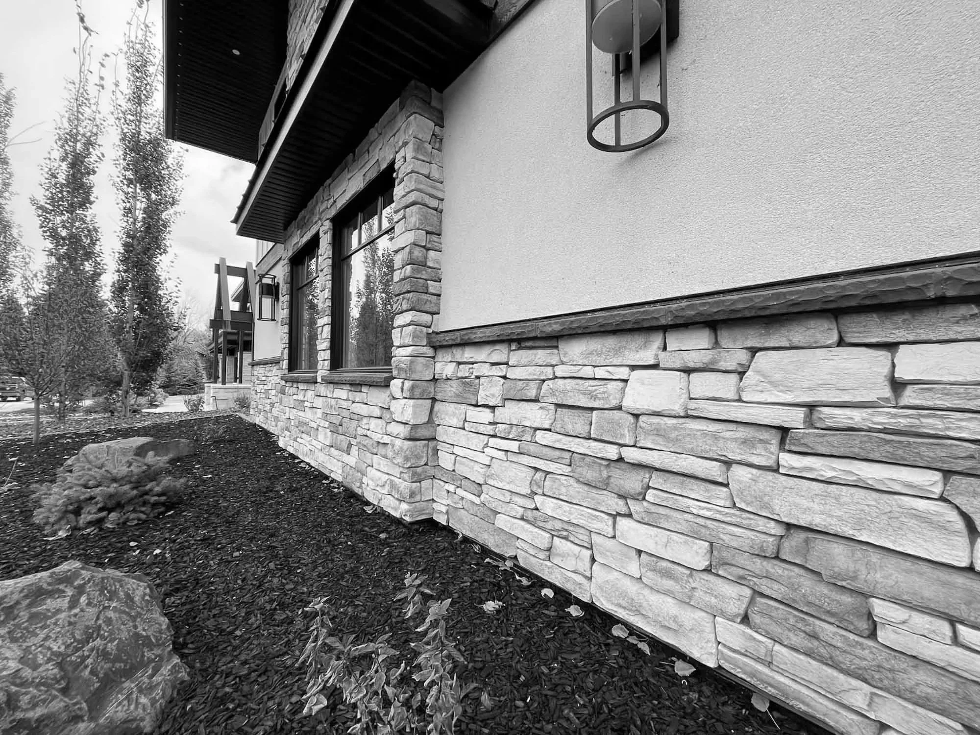 Black and white photo of the exterior of a house with stone and stucco walls, window, and a wall-mounted outdoor light fixture. There are trees and a garden in the background.