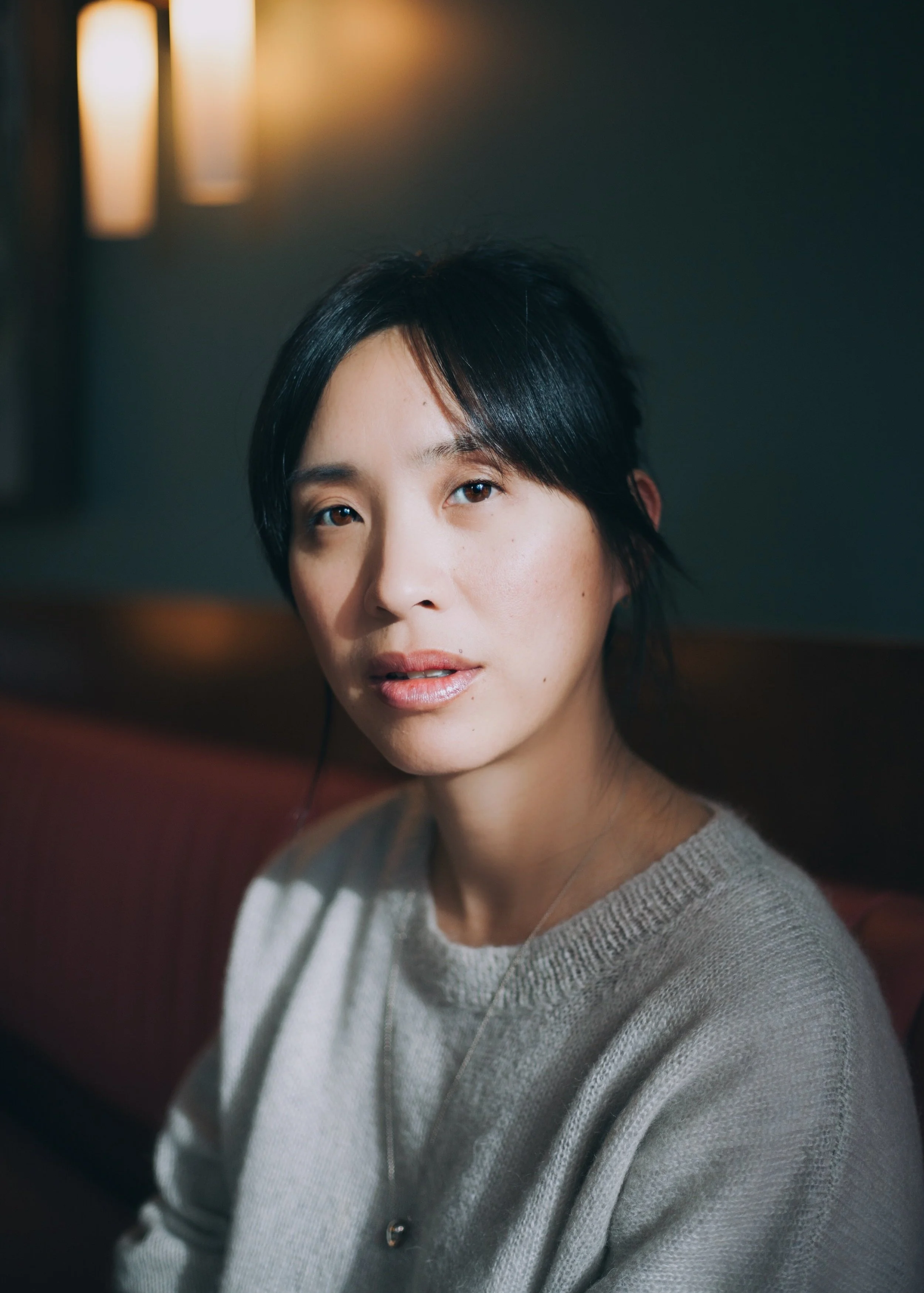 Woman sitting indoors with soft focus lighting, wearing a grey sweater.