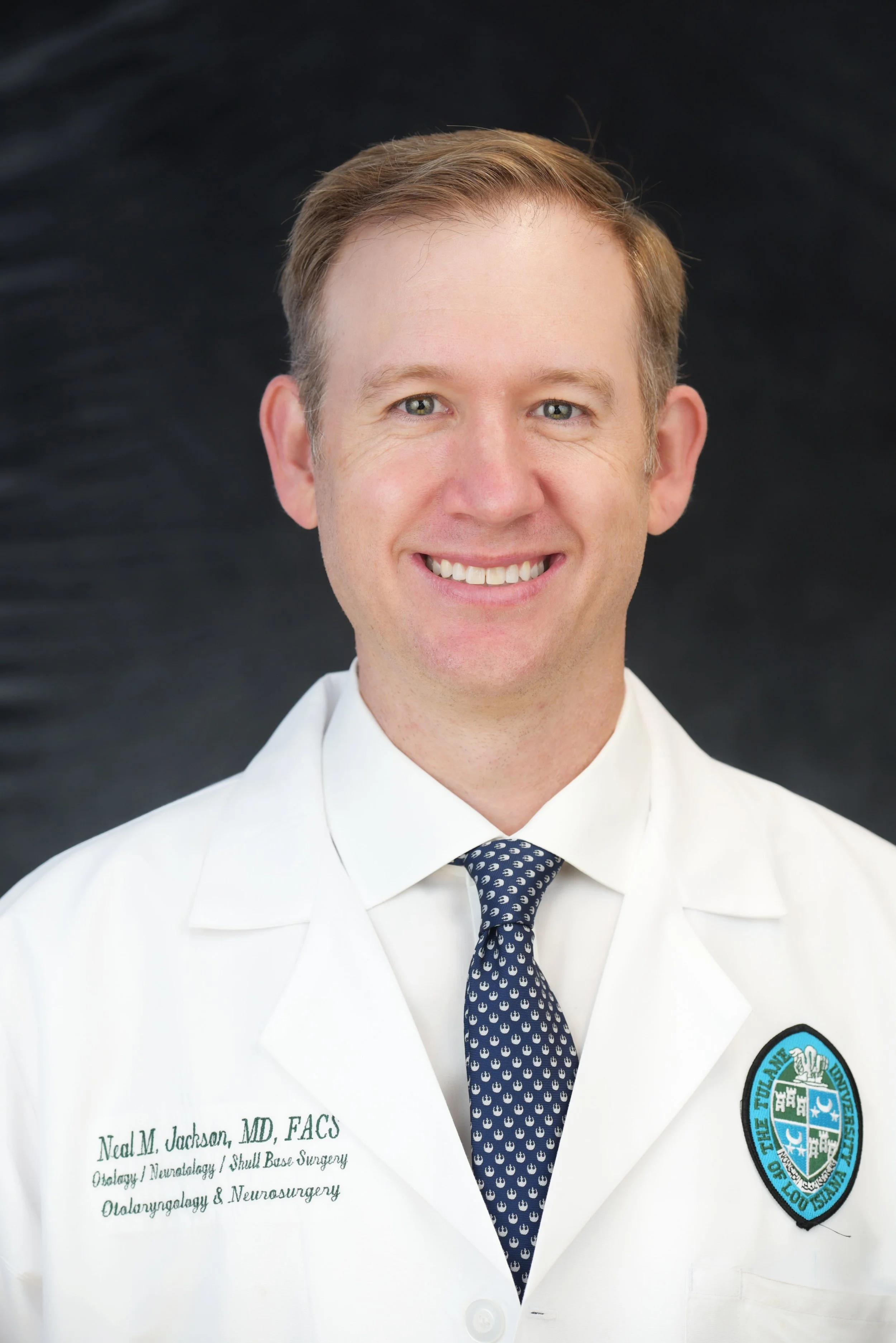 A smiling man in a white medical coat with Tulane University patch, wearing a dark patterned tie, standing against a dark background.