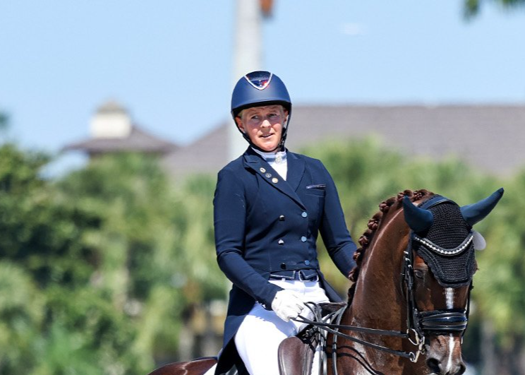 A woman in equestrian attire riding a horse, wearing a helmet and gloves, outdoors on a sunny day.