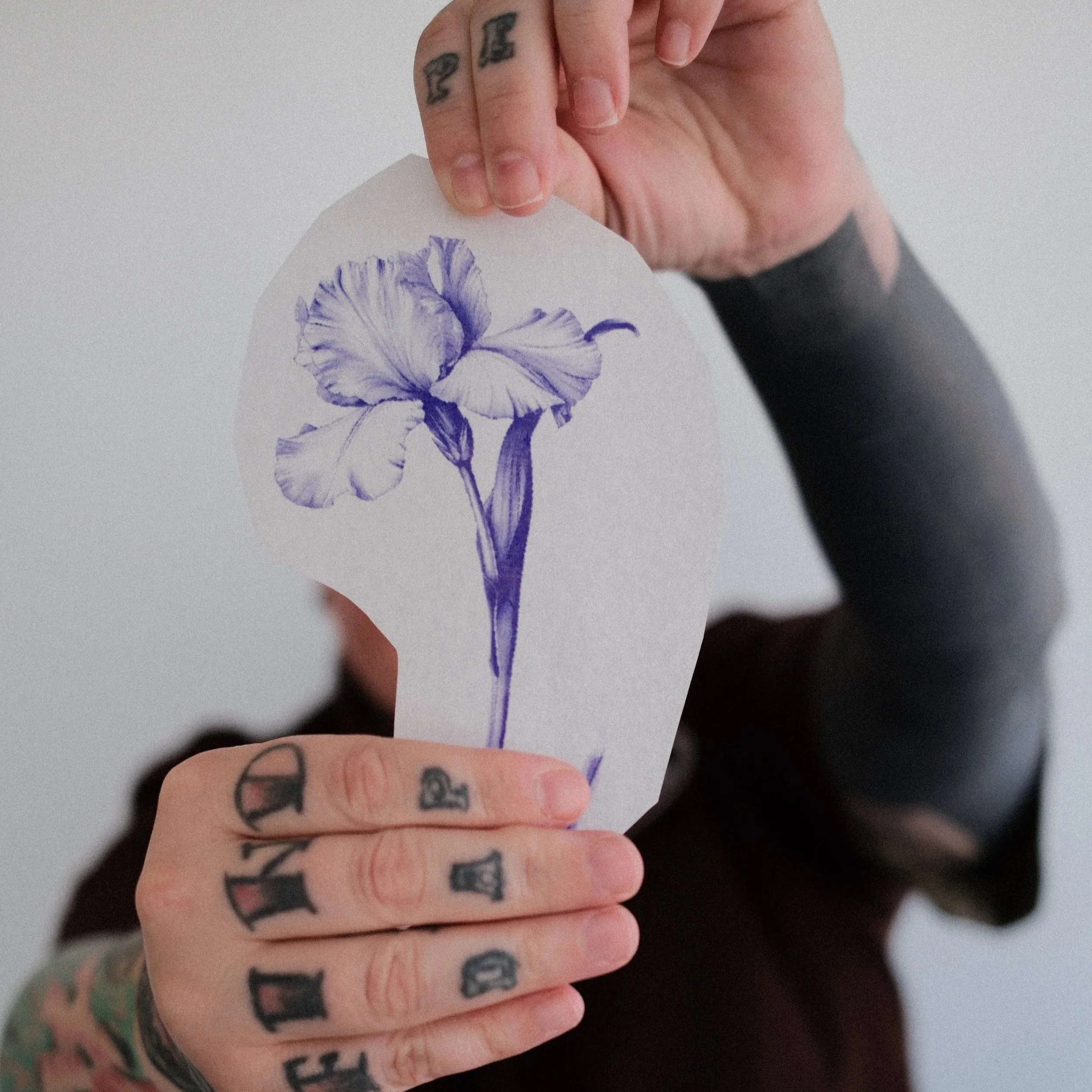 Minnesota tattoo studio owner, Ryane Rose holding an iris flower tattoo stencil, with visible tattoos on fingers and hand.