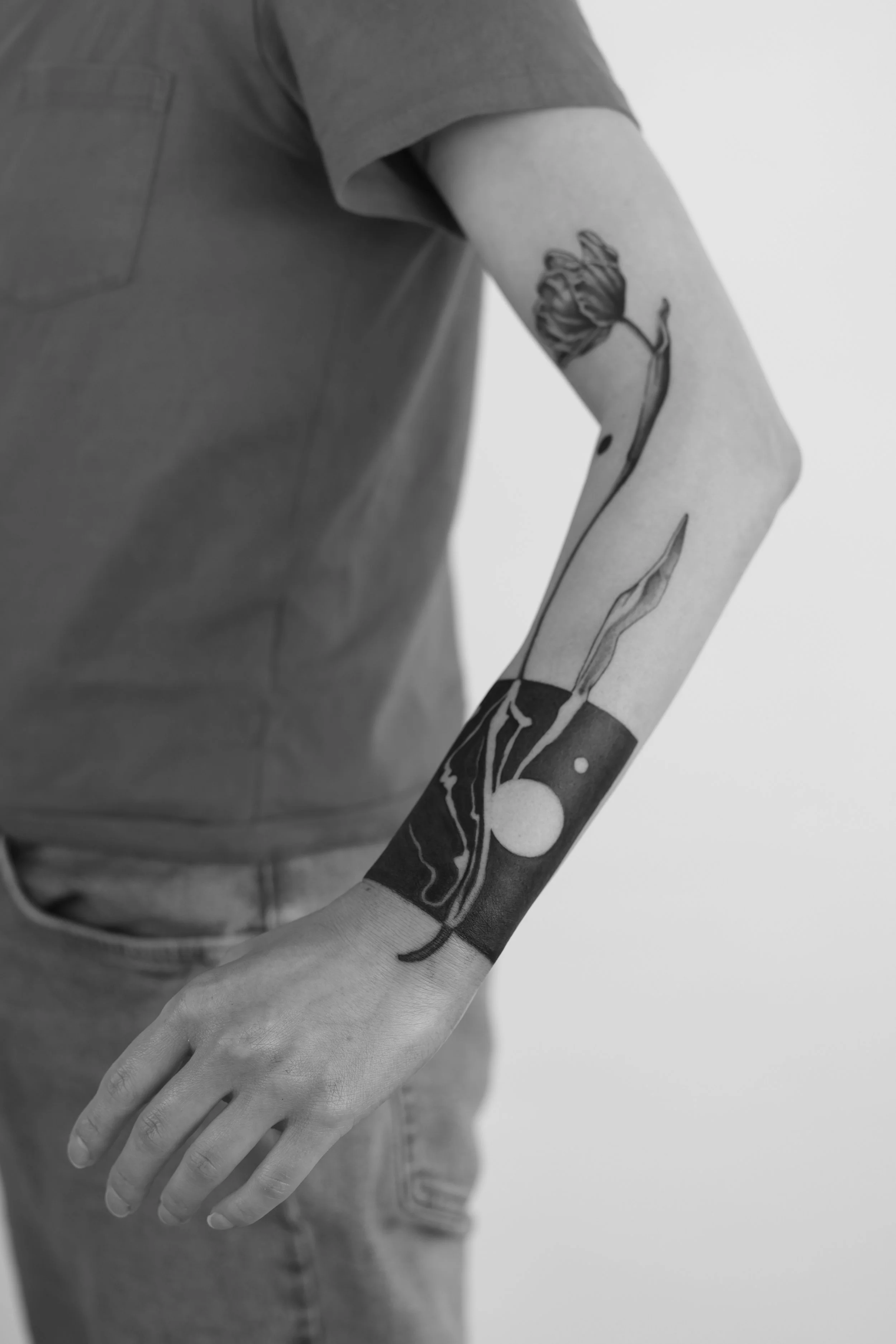A person with a floral sleeve tattoo on their arm, wearing a gray T-shirt and jeans, standing with their hand relaxed by their side.