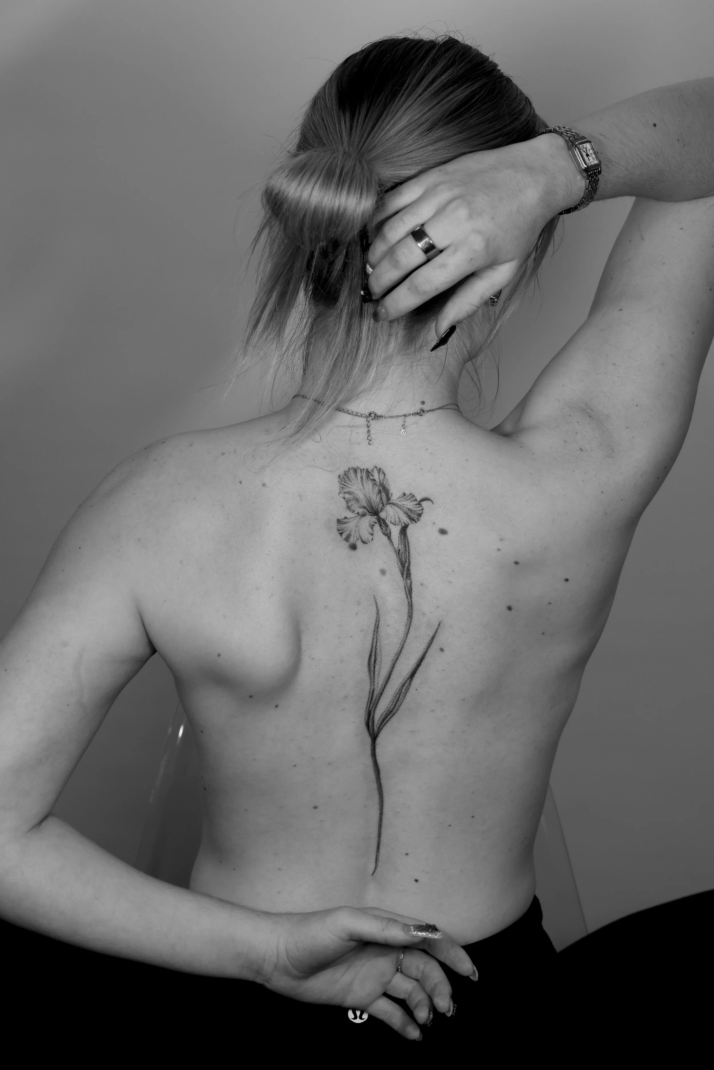 A person with light skin and long hair tied in a bun is posing topless against a plain background. They have a floral tattoo on their back, a necklace, rings, and a bracelet. Their right arm is raised, and their left hand is resting on their hip.