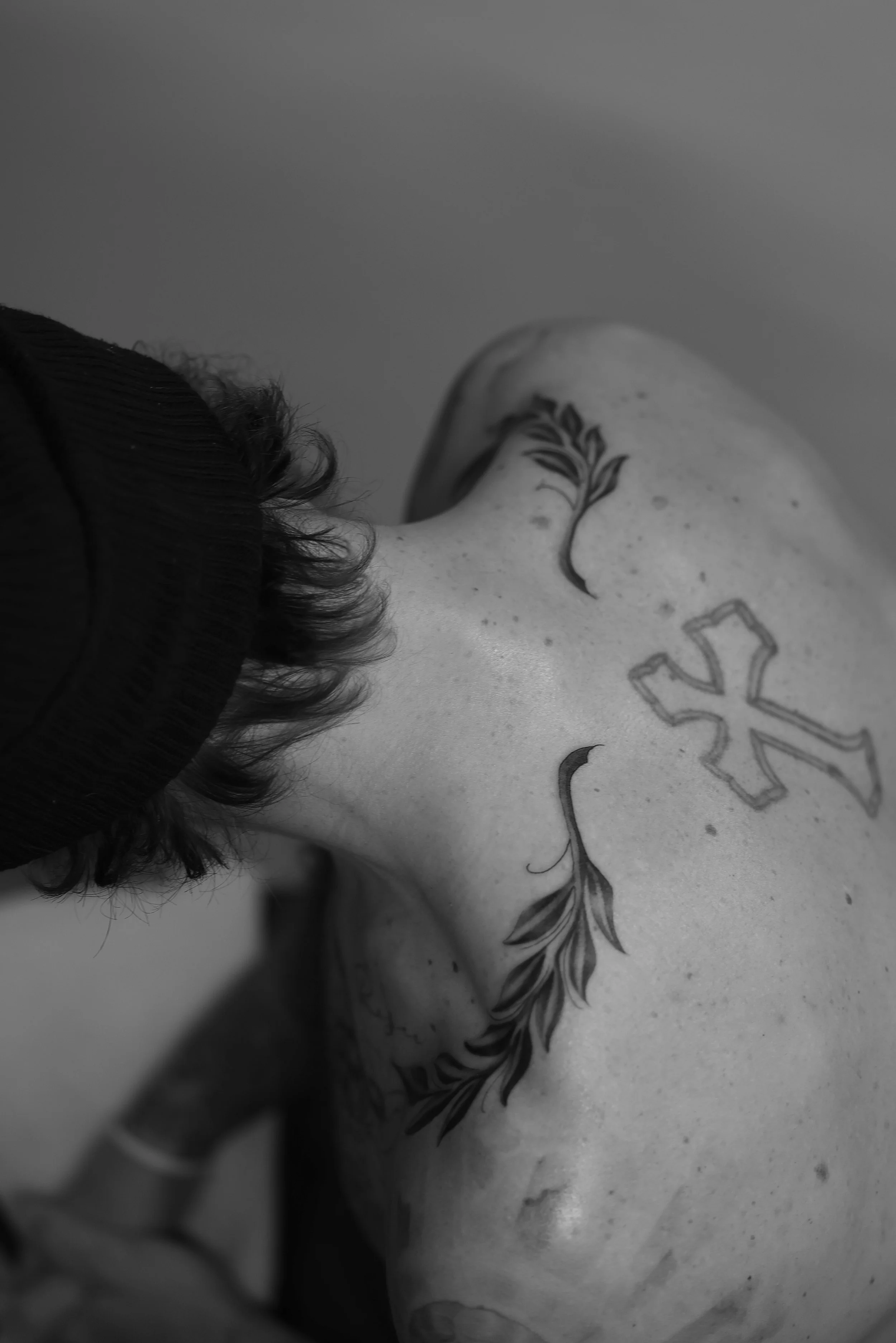 Close-up of a person's back with tattoos, including a branch with leaves and a geometric design, and part of the person's curly hair and clothing visible.