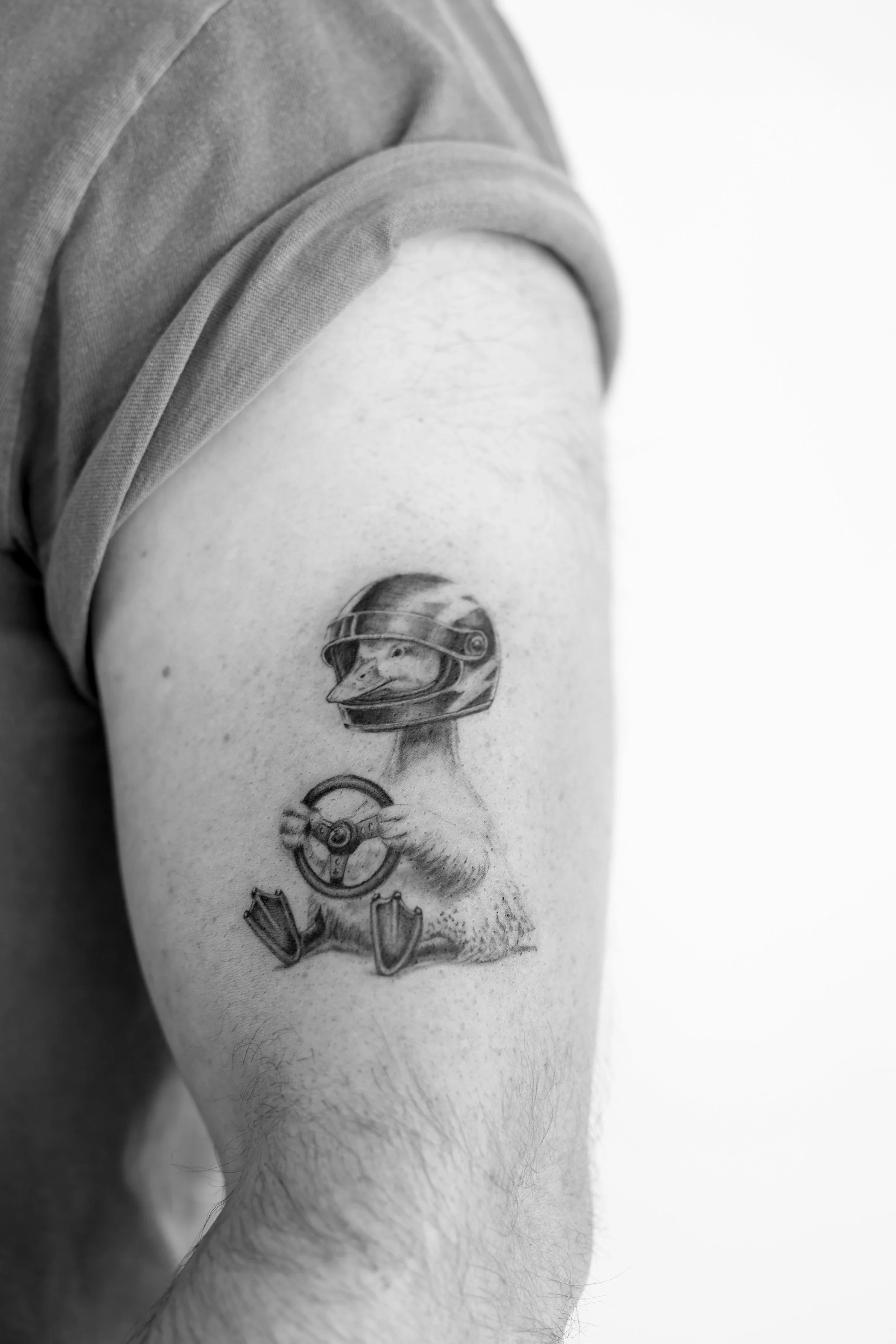 Close-up black and white photo of a person's upper arm with a tattoo of a duck wearing a racing helmet. The person is wearing a rolled-up T-shirt.