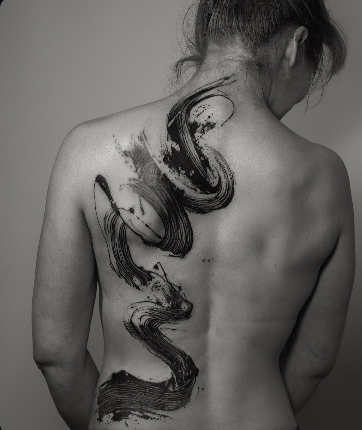 A person with long hair tied back is turned to the side, showing their back and shoulder. They have an artistic black tattoo with abstract flowing lines and splashes that covers a large part of their back and shoulder.