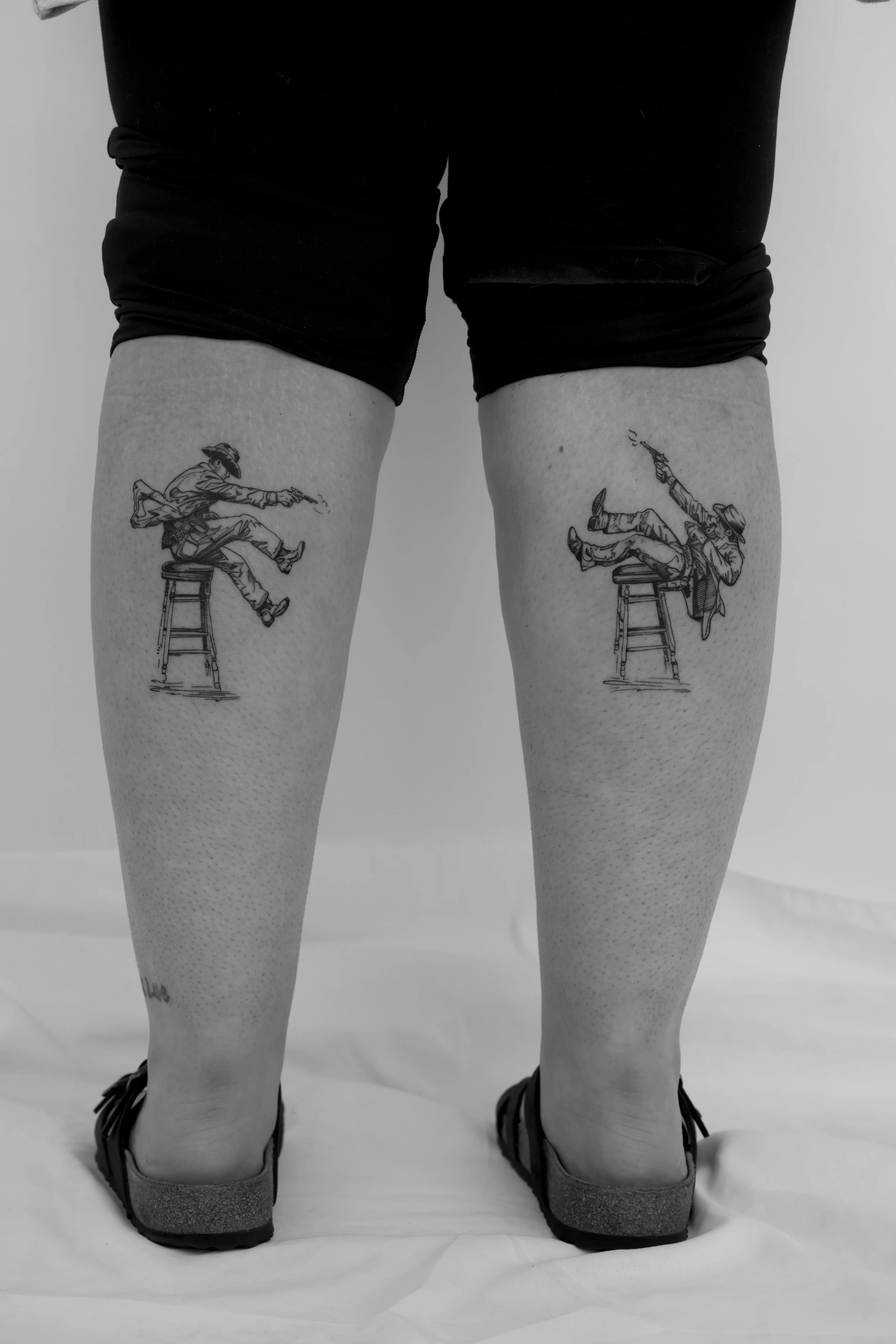 Person wearing shorts with tattoos on thighs, standing on a white surface, wearing shoes. The image is in black and white.
