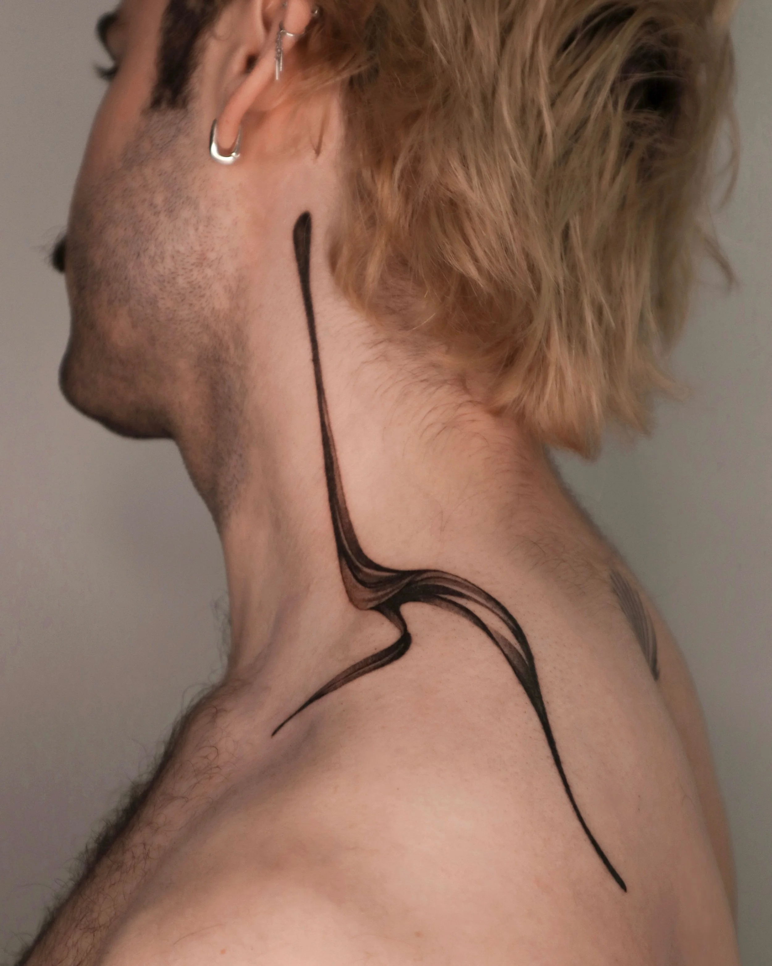 Close-up of a person's neck and side of face with blond, tousled hair, wearing multiple earrings, and a black tattoo on the neck.