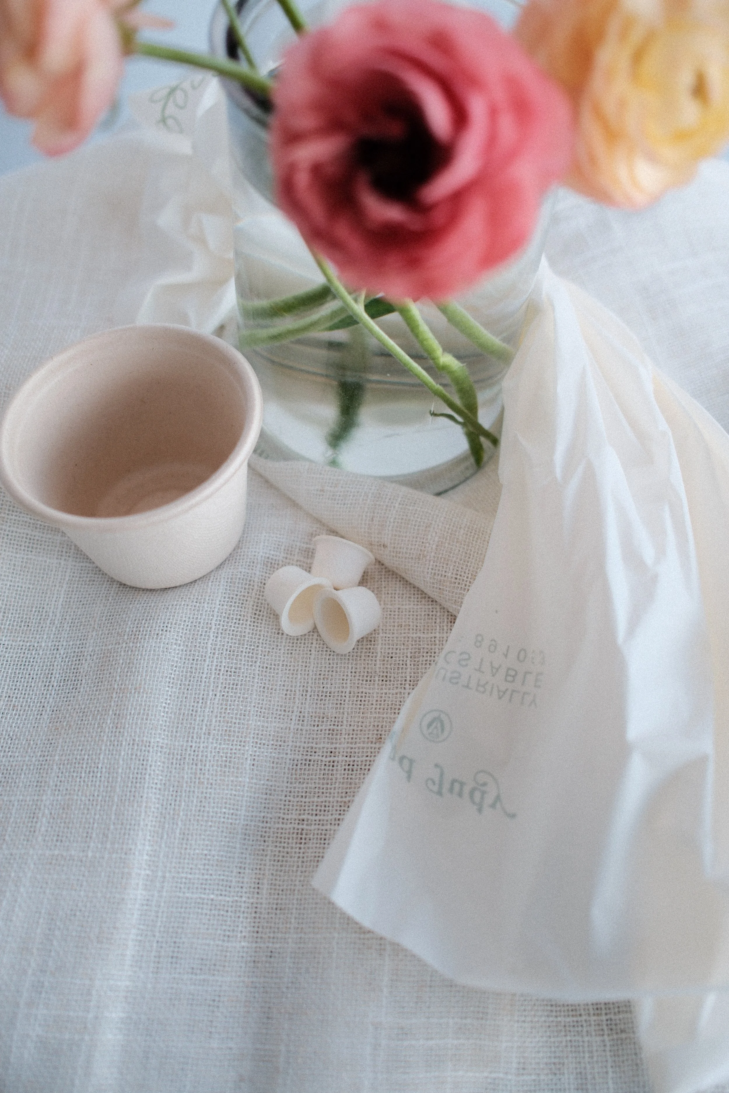 A white tablecloth with a clear glass vase holding pink, peach, and yellow ranunculus flowers, a white cup with a hot beverage, small white ceramic cups, and a partially visible paper or card with printed text.