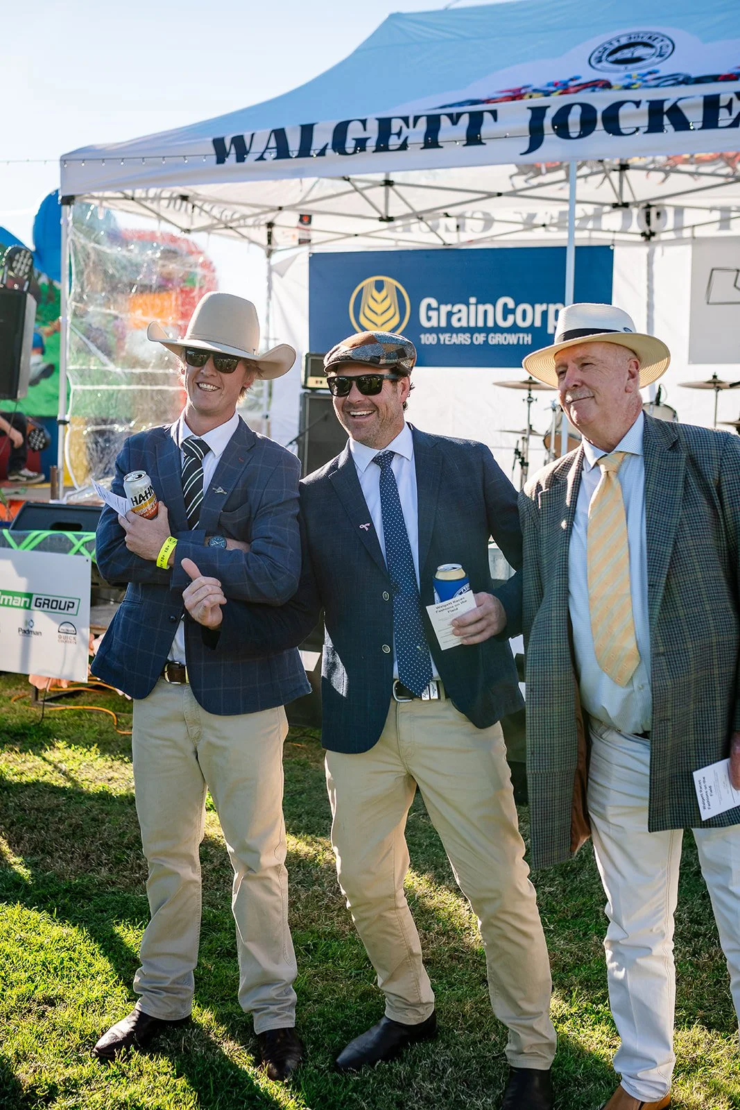 Three men in suits and hats participating in the Fashion on the field at the Walgett Races, smiling and holding drinks, with a tent and signage behind them.