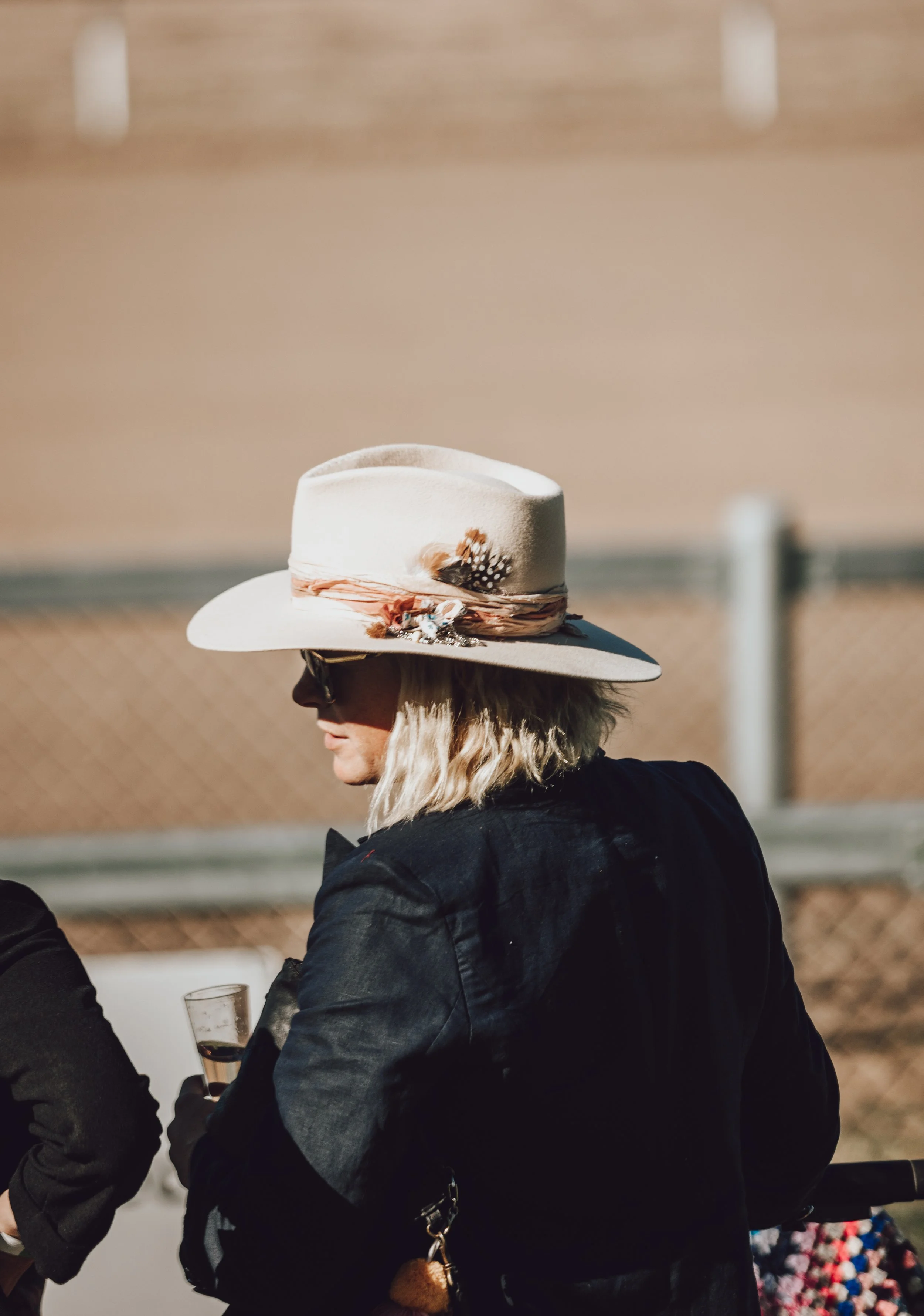 A person wearing a wide-brimmed white hat with decorative adornments and feathers, sunglasses, and a dark jacket, holding a drink in their hand.