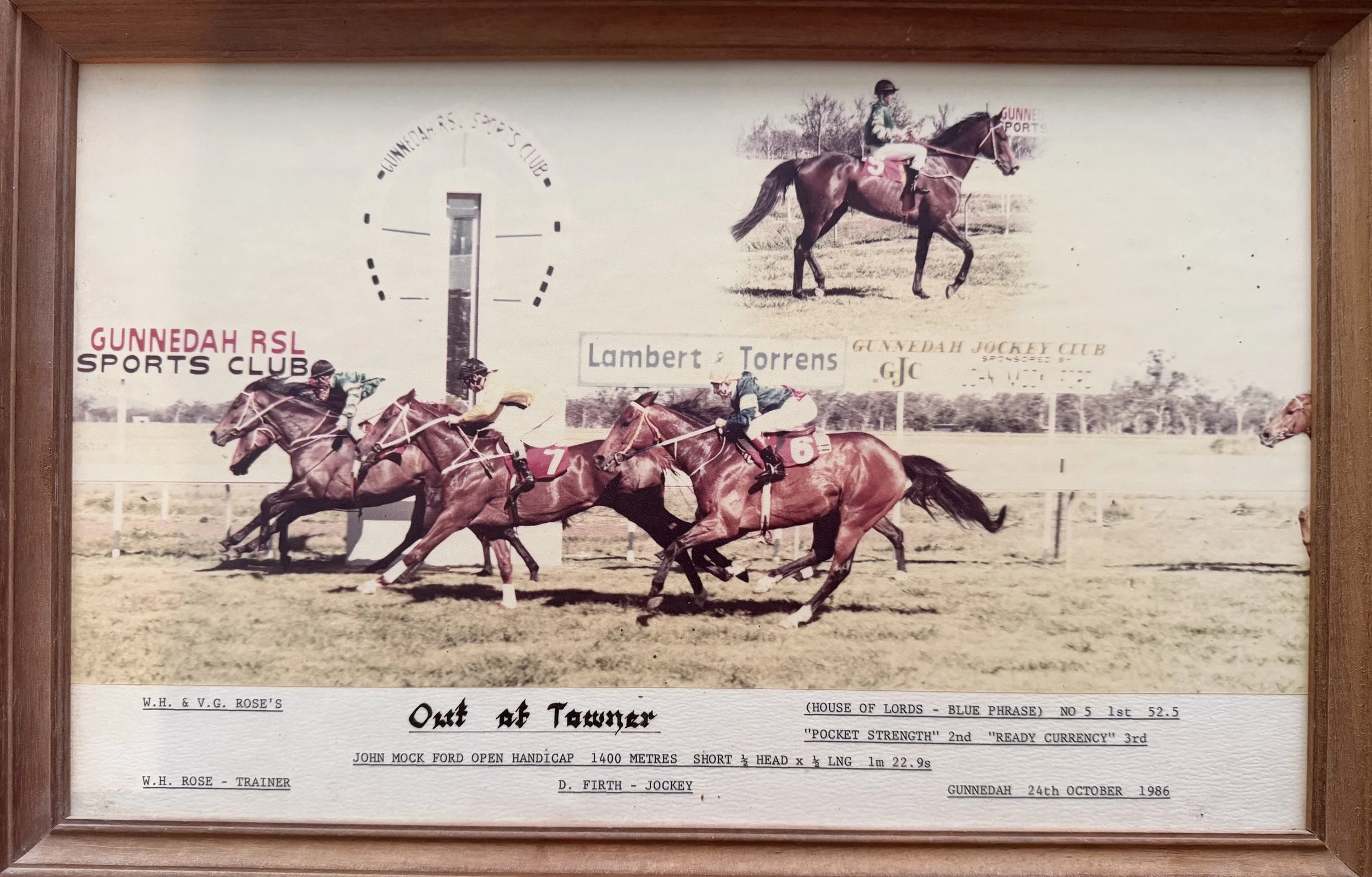 Photograph of a horse racing event at Gunnedah Races 1986, with multiple horses and jockeys in race, set in a grassy field with advertising signs and a scoreboard in the background. Jockey: Chris Troy