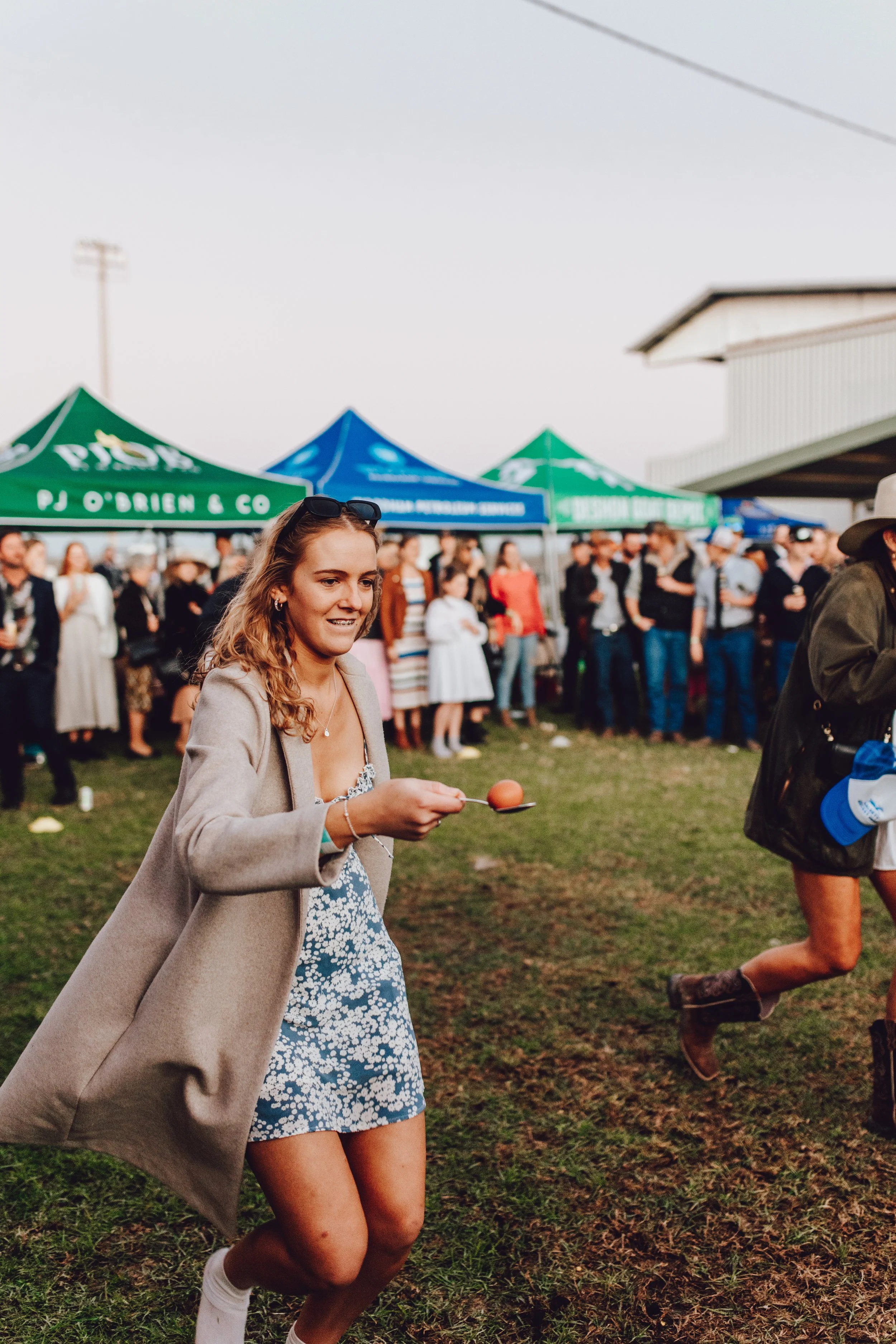 Young woman with wavy hair, sunglasses on her head, wearing a light-colored blazer and a floral dress, holding an egg on a spoon while walking on a grassy field during a daytime outdoor event with tents in the background and a crowd of people.
