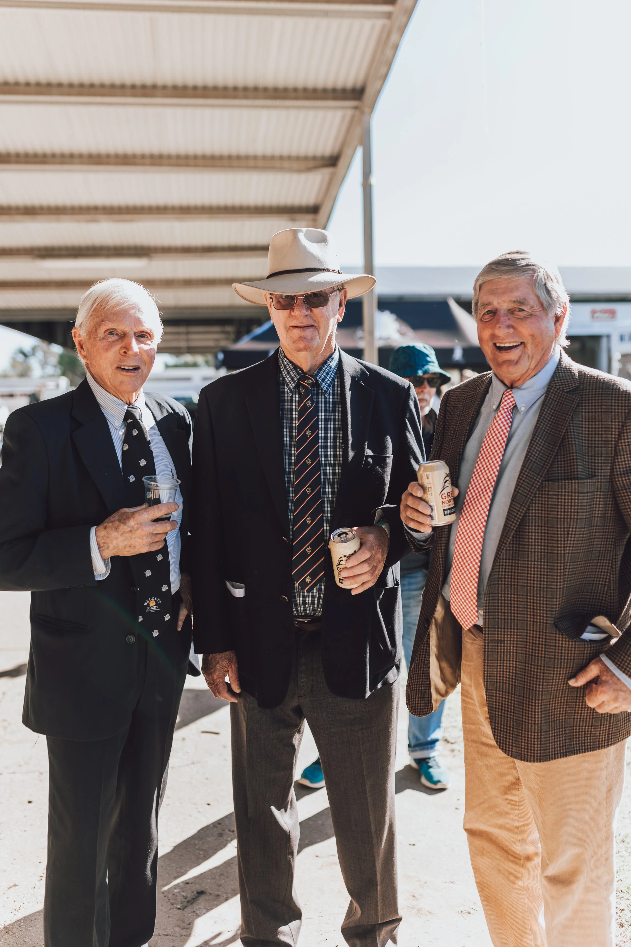 Three elderly men dressed in suits and ties, holding drinks, smiling outdoors on a sunny day at the Walgett races.