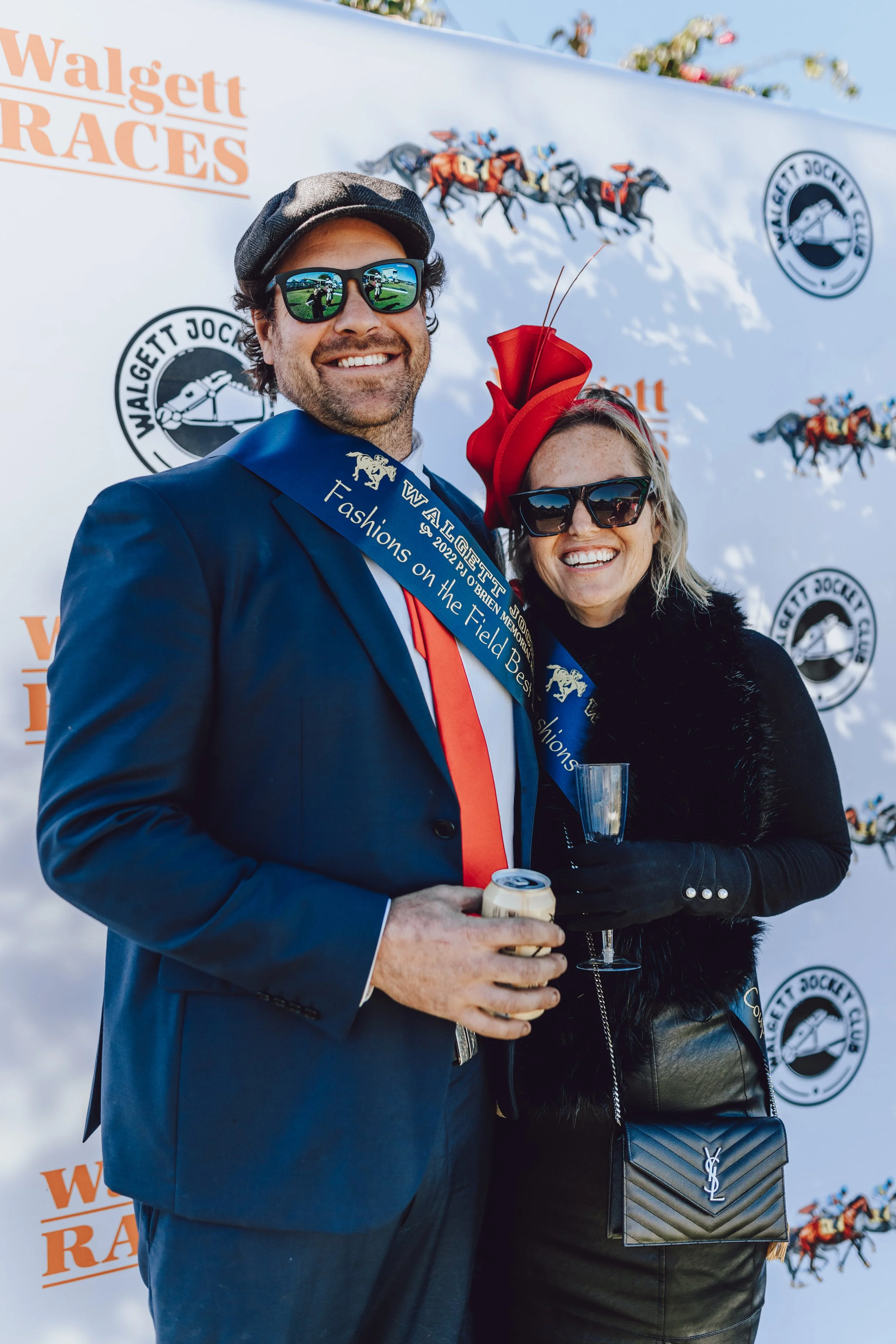 Two people dressed in formal attire, smiling, at a racetrack event with Walgett Races and Walgett Jockey Club banners in the background. The man wears sunglasses, a suit, and a hat, and has a sash "Fashions on the Field " at the Walgett Races.