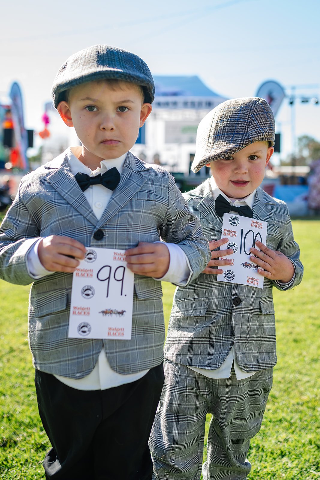 Two young boys dressed in vintage-style suits and bow ties, holding race bibs with the numbers 99 and 100, standing outdoors at a race event with a stage and tents in the background. fashion on the field at the Walgett Races.