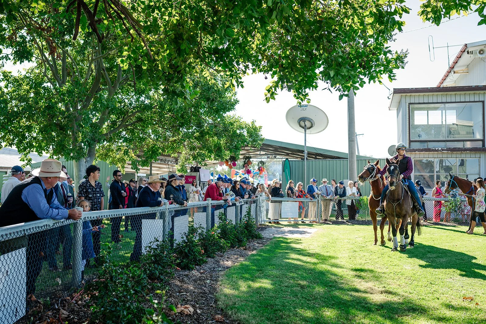 Walgett Races Dress Circle | People watching a horse riding show outdoors, with some riders on horseback and an audience behind a fence under trees on a sunny day.