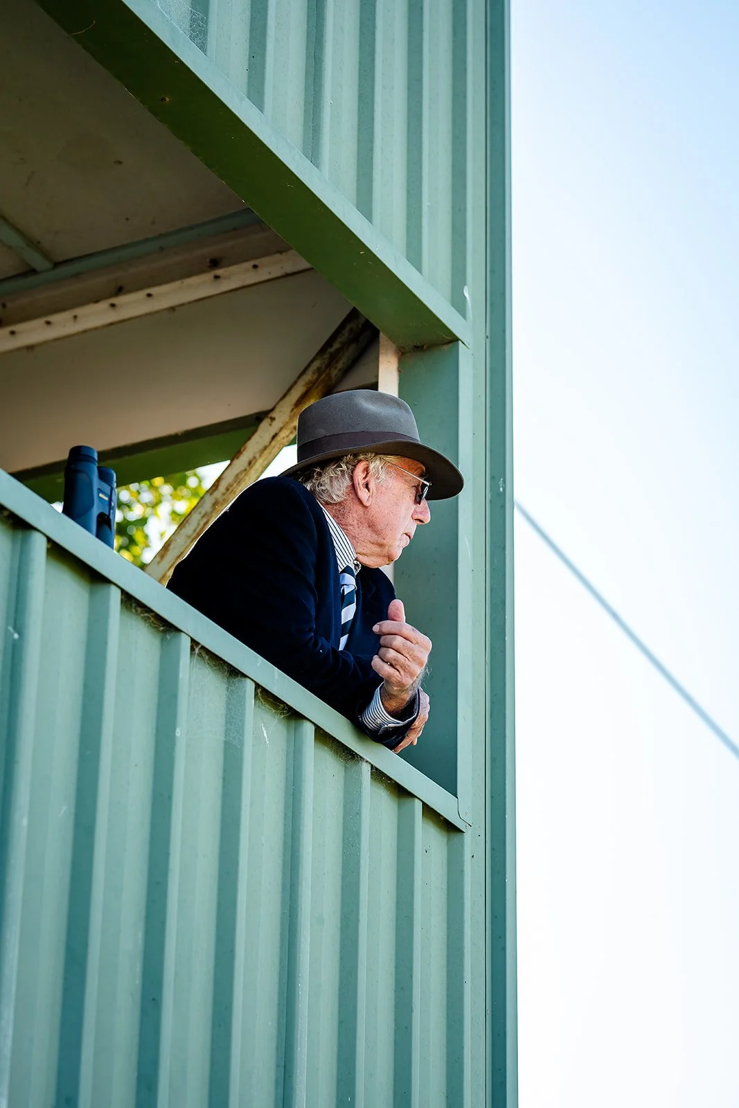 An older man in a suit, sunglasses, and a gray hat leaning on a green balcony country racing railing, looking out.