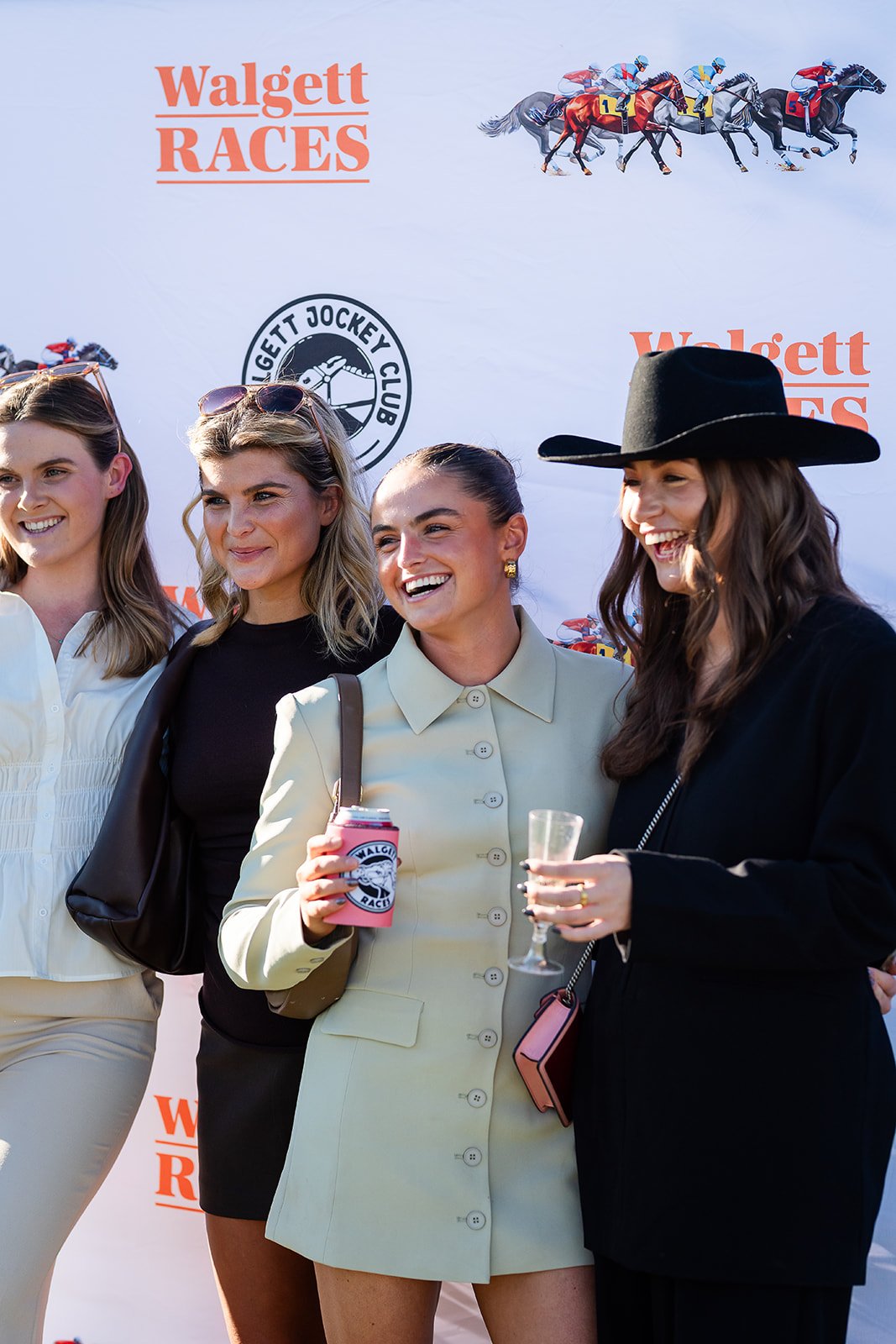 Four women standing together at the Walgett Races, smiling, with a backdrop featuring horses racing and logos for Walgett Races and Walgett Jockey Club.